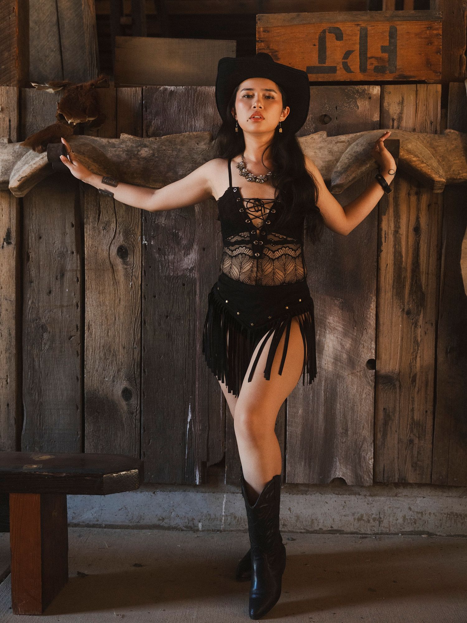 Woman in black western outfit with cowboy hat and boots posing in rustic barn.