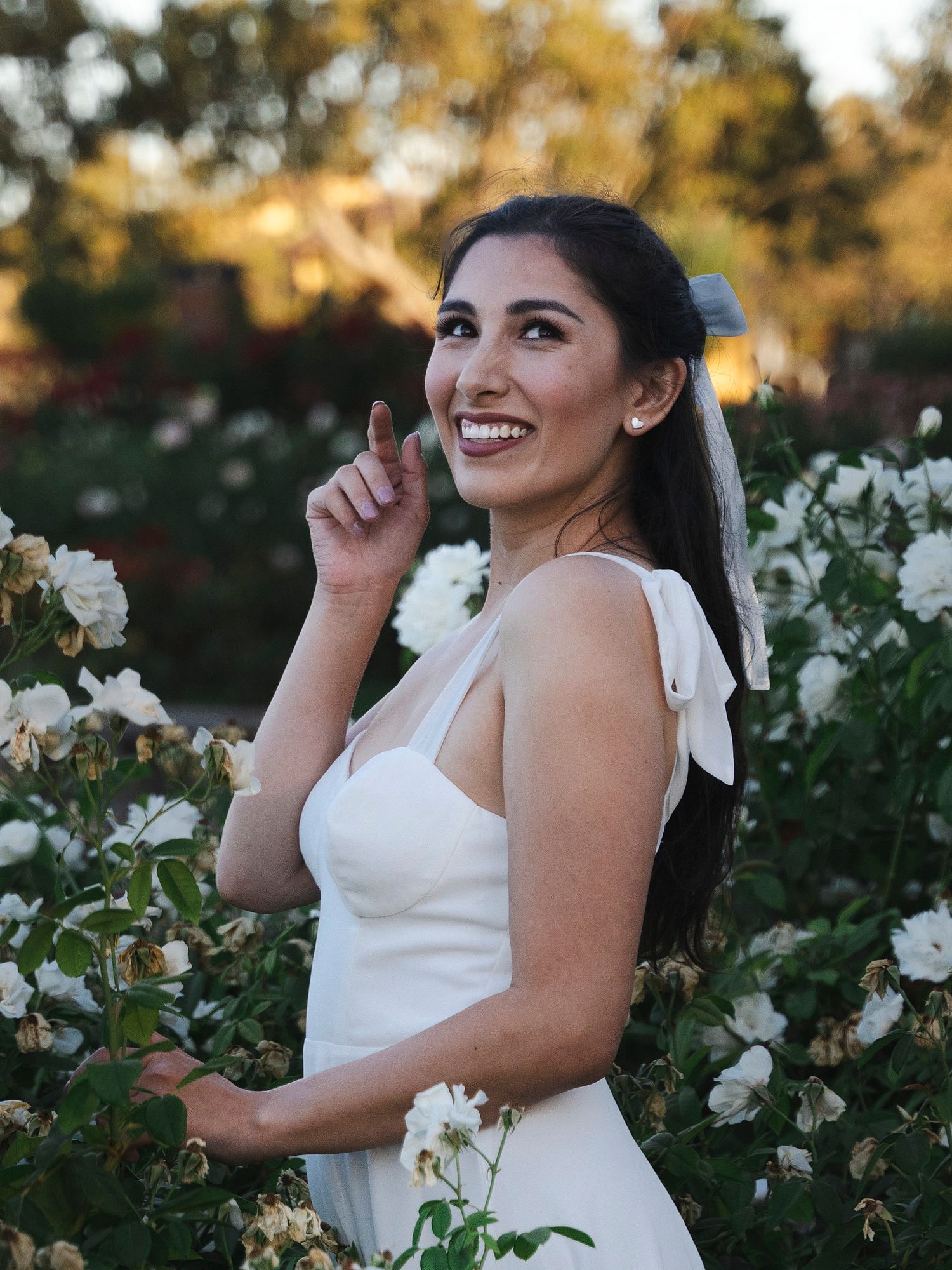 A smiling woman in a white dress stands among blooming white roses in a garden at sunset.