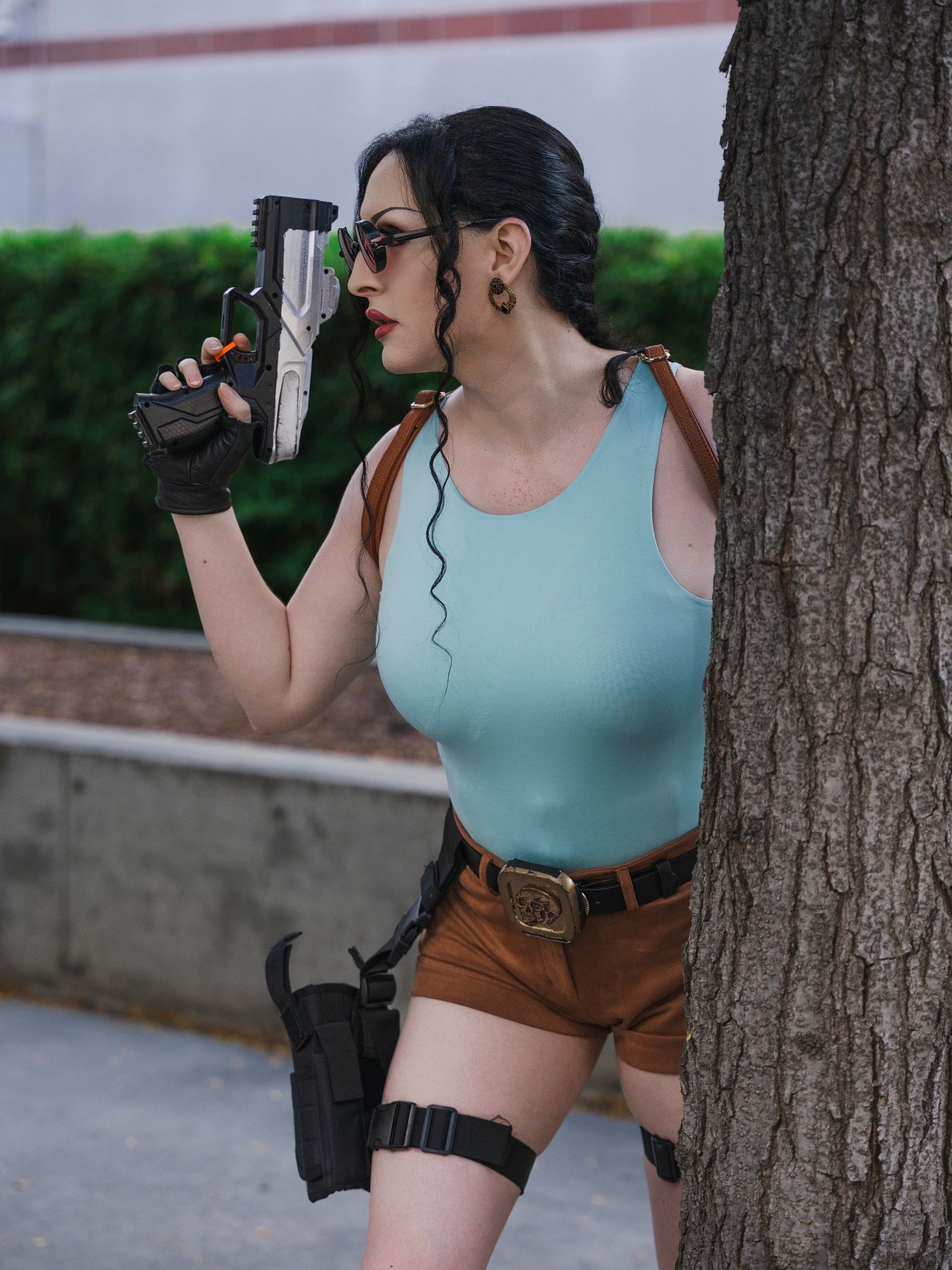 Woman in Lara Croft cosplay costume holding prop gun beside a tree outdoors.