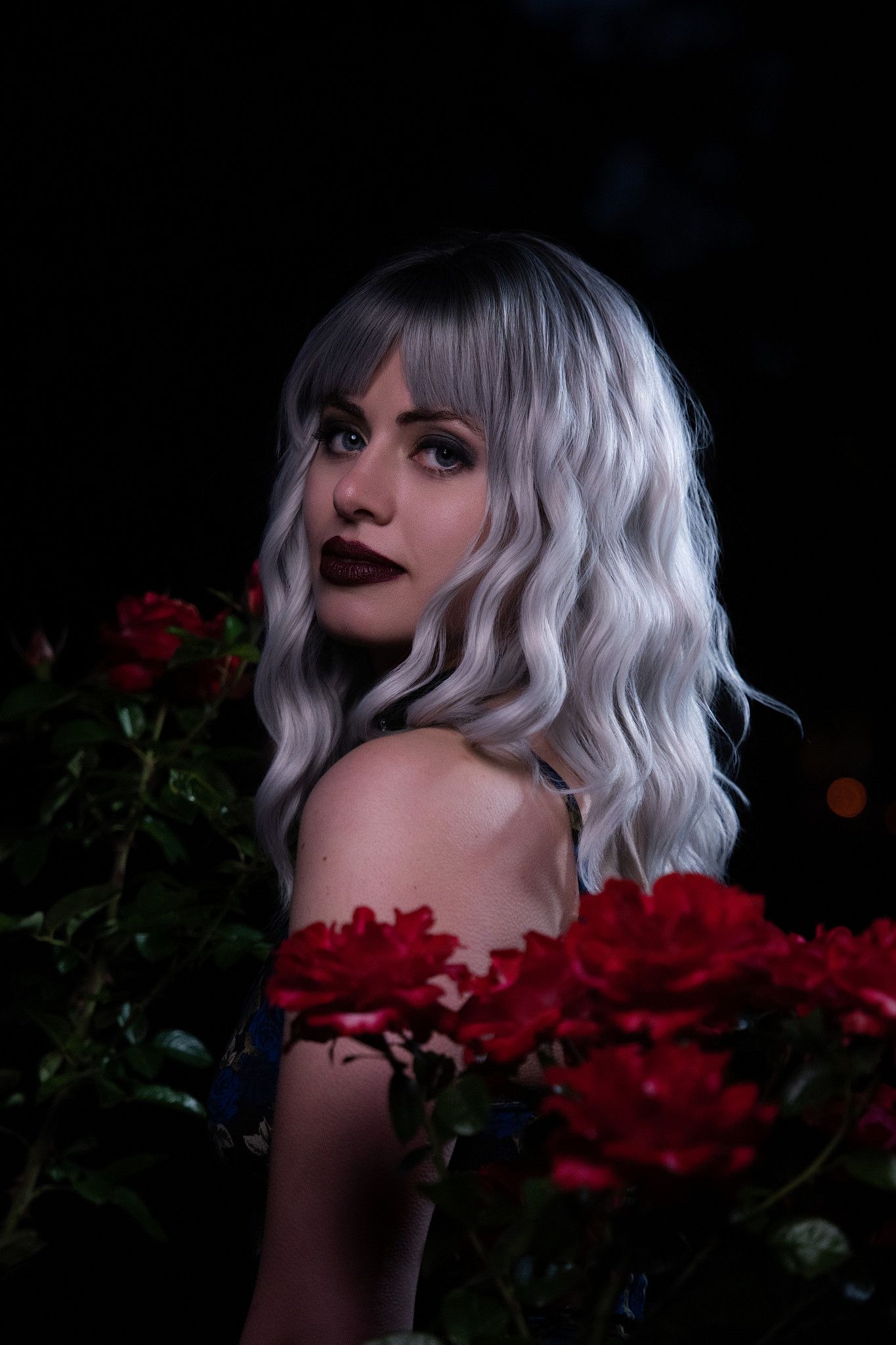 Silver-haired woman with bold dark lips surrounded by vibrant red roses in darkness.