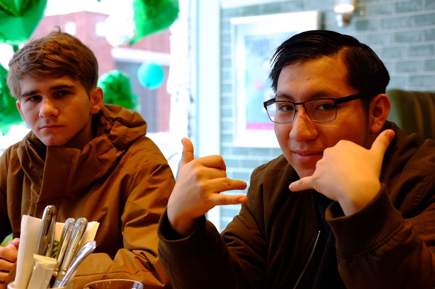 Two young men in brown jackets sitting at a table, one making a hand gesture, green decor behind.