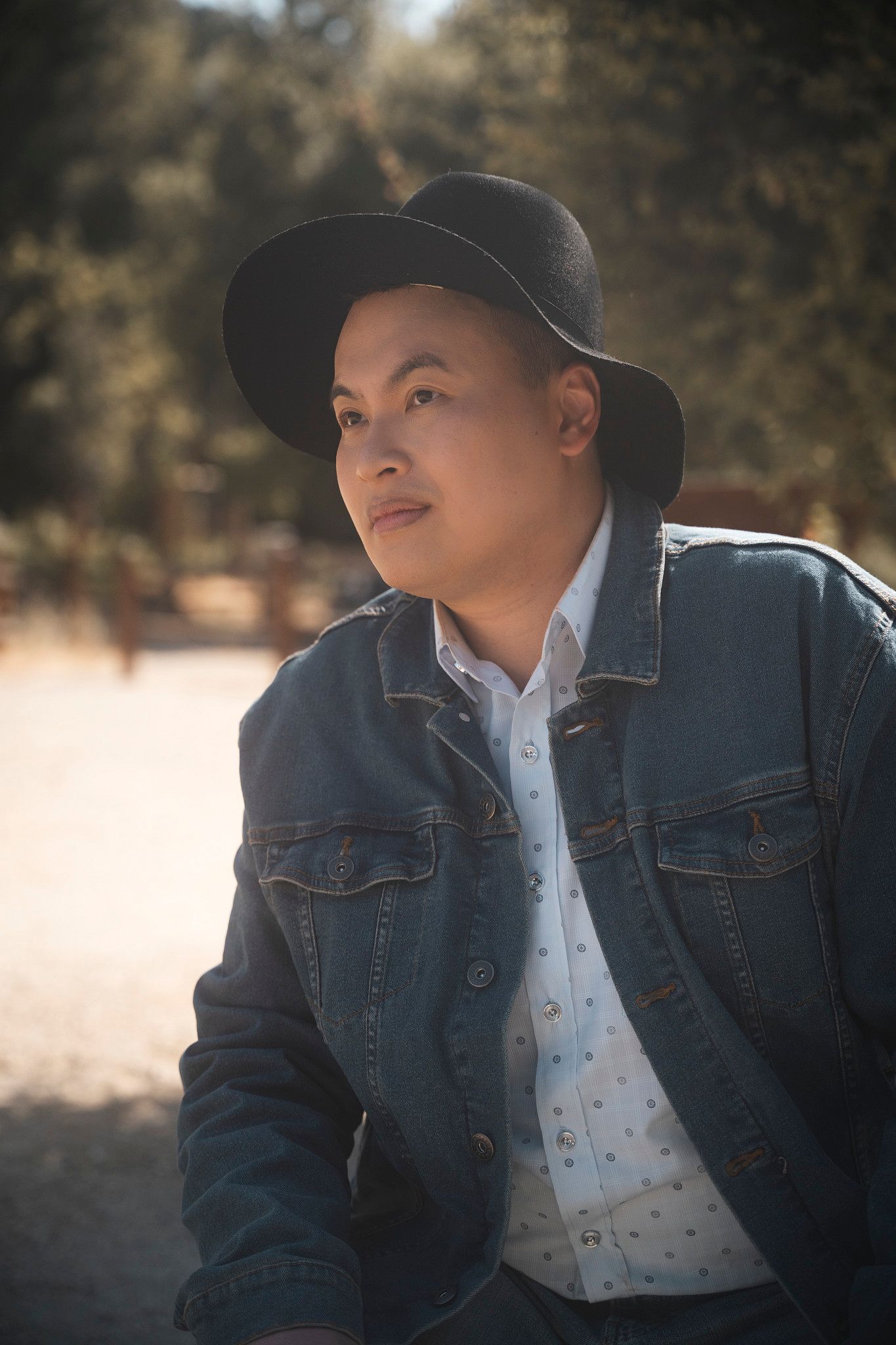 Person wearing black hat and denim jacket sitting outdoors in natural sunlight.