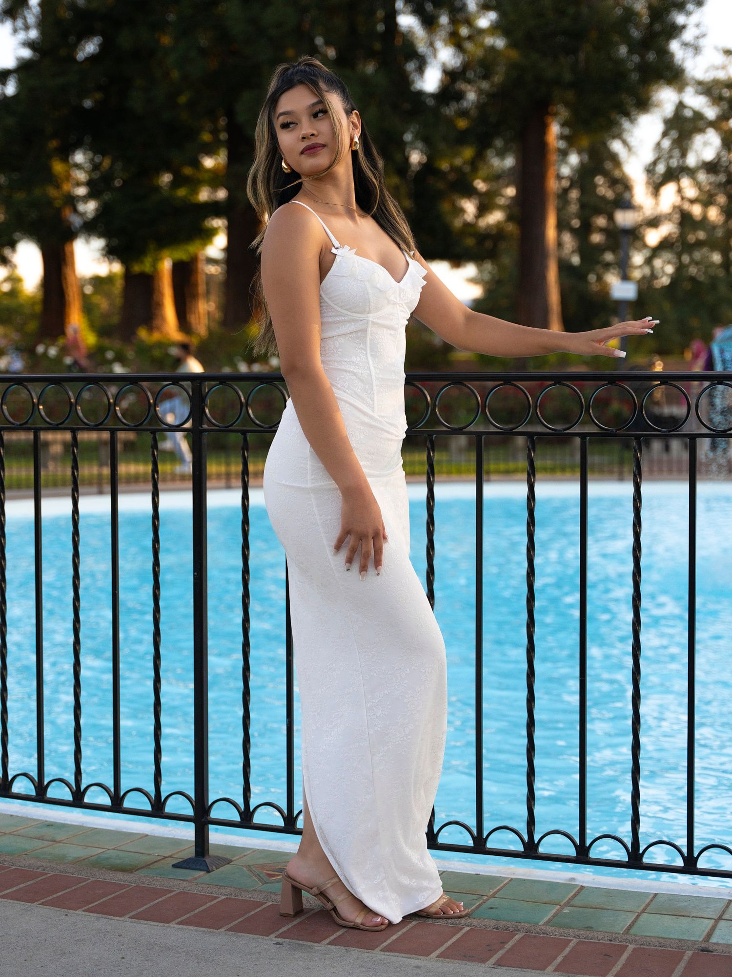 Woman in white maxi dress posing by pool with iron fence and trees in background.