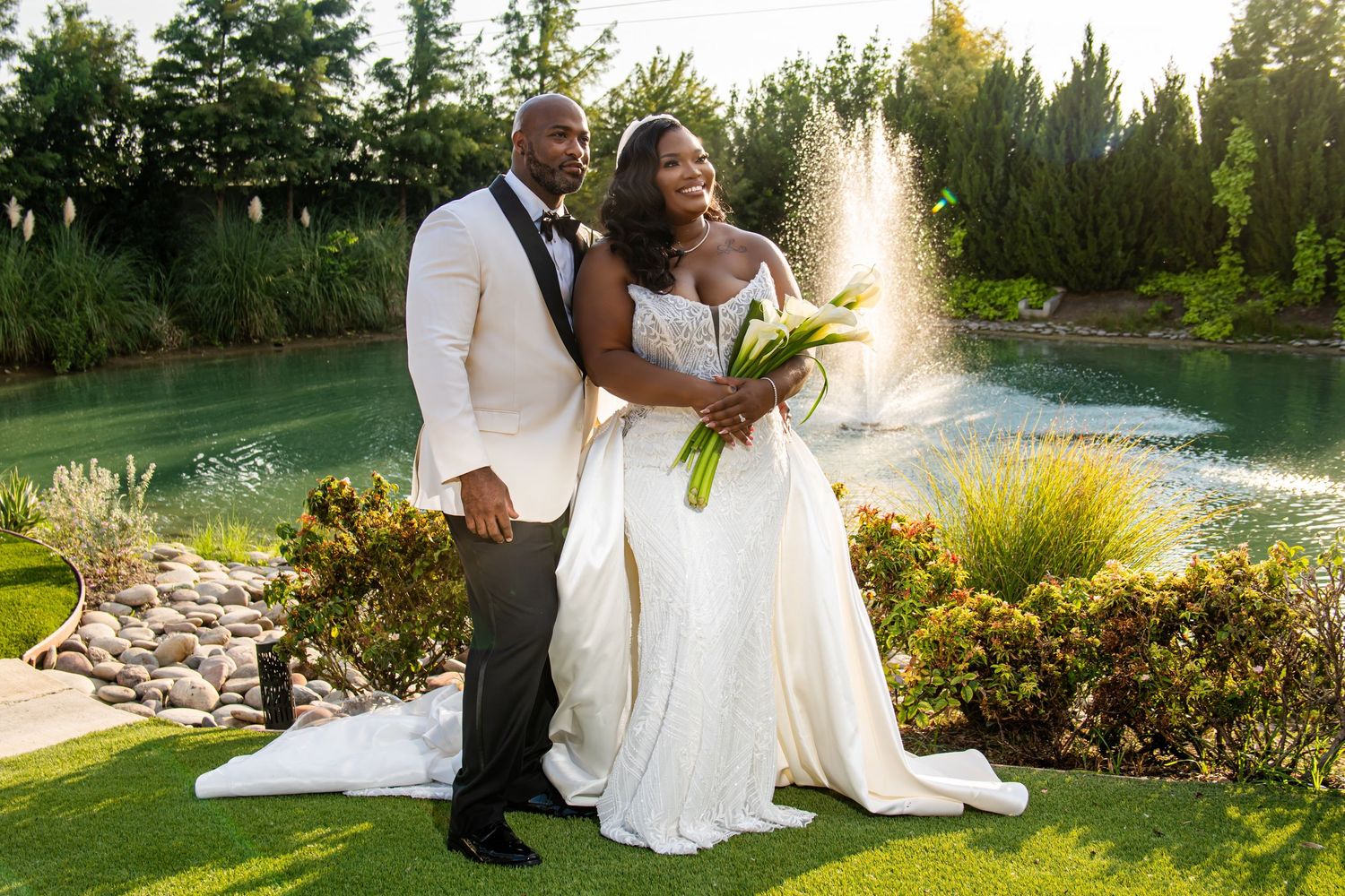 A newlywed couple stands together on manicured grounds with a fountain in the background.