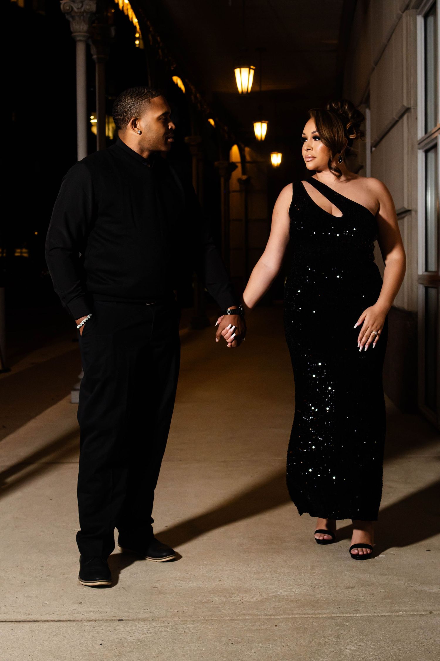 Stylish couple in formal attire poses in dimly lit hallway with vintage lamps.