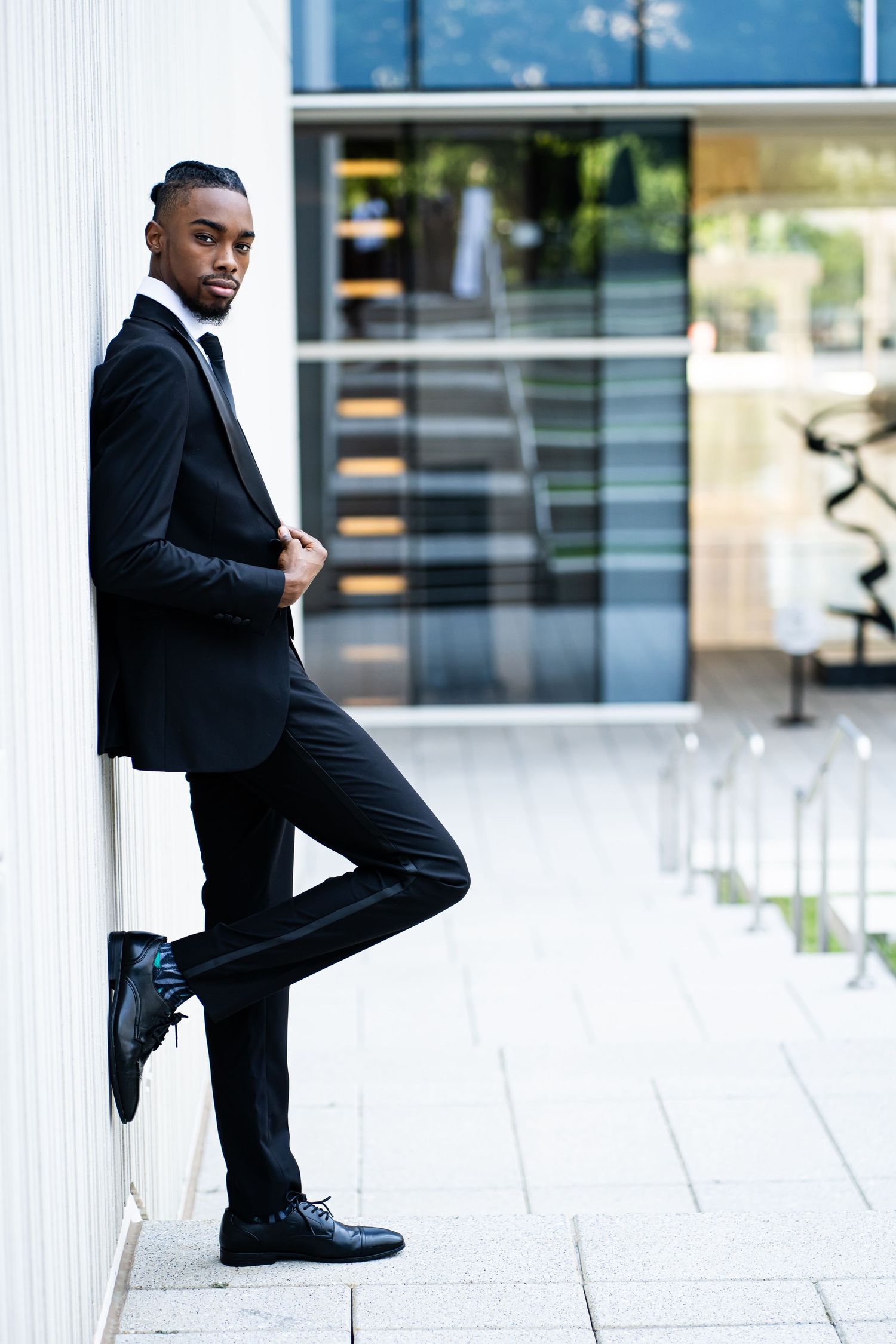 Groom poses confidently in classic black tuxedo against modern white architectural backdrop.