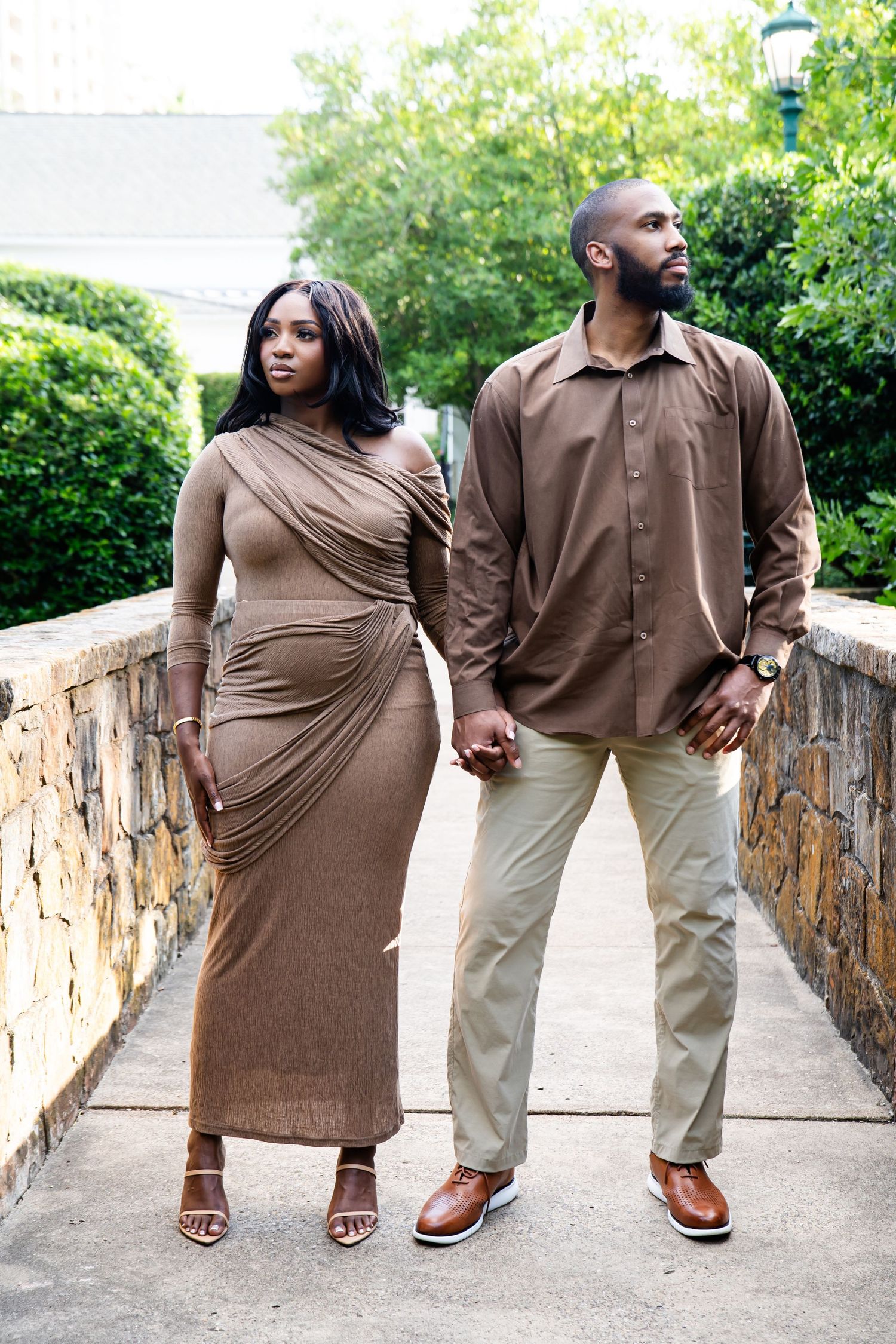 Couple in coordinating brown outfits pose together on a stone pathway surrounded by greenery.