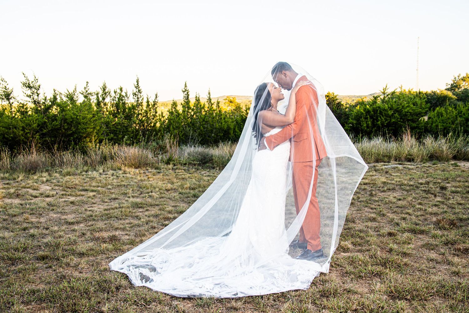 Bride and groom posing with flowing veil in outdoor field at sunset.