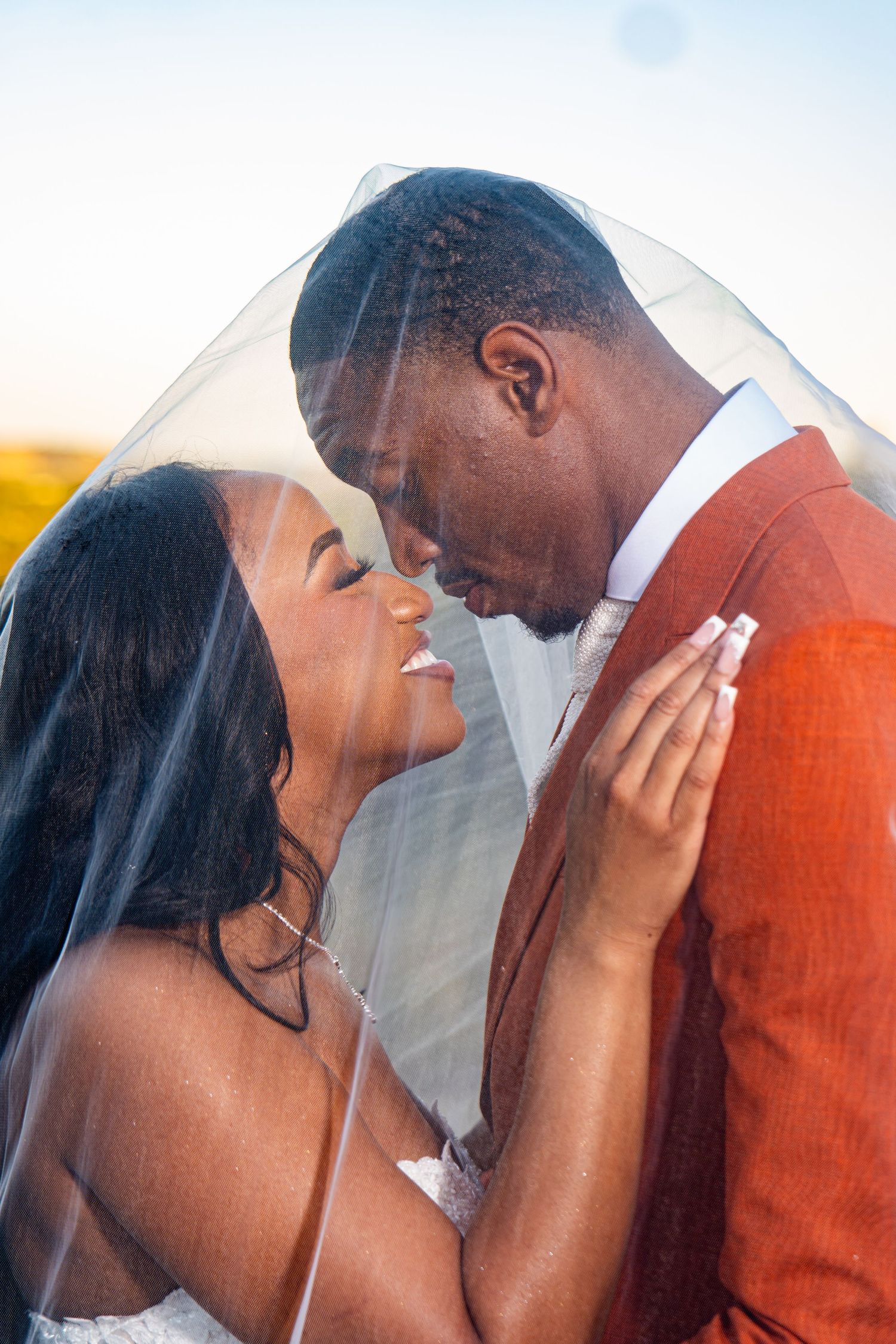 Romantic wedding couple embracing under veil during sunset photoshoot.