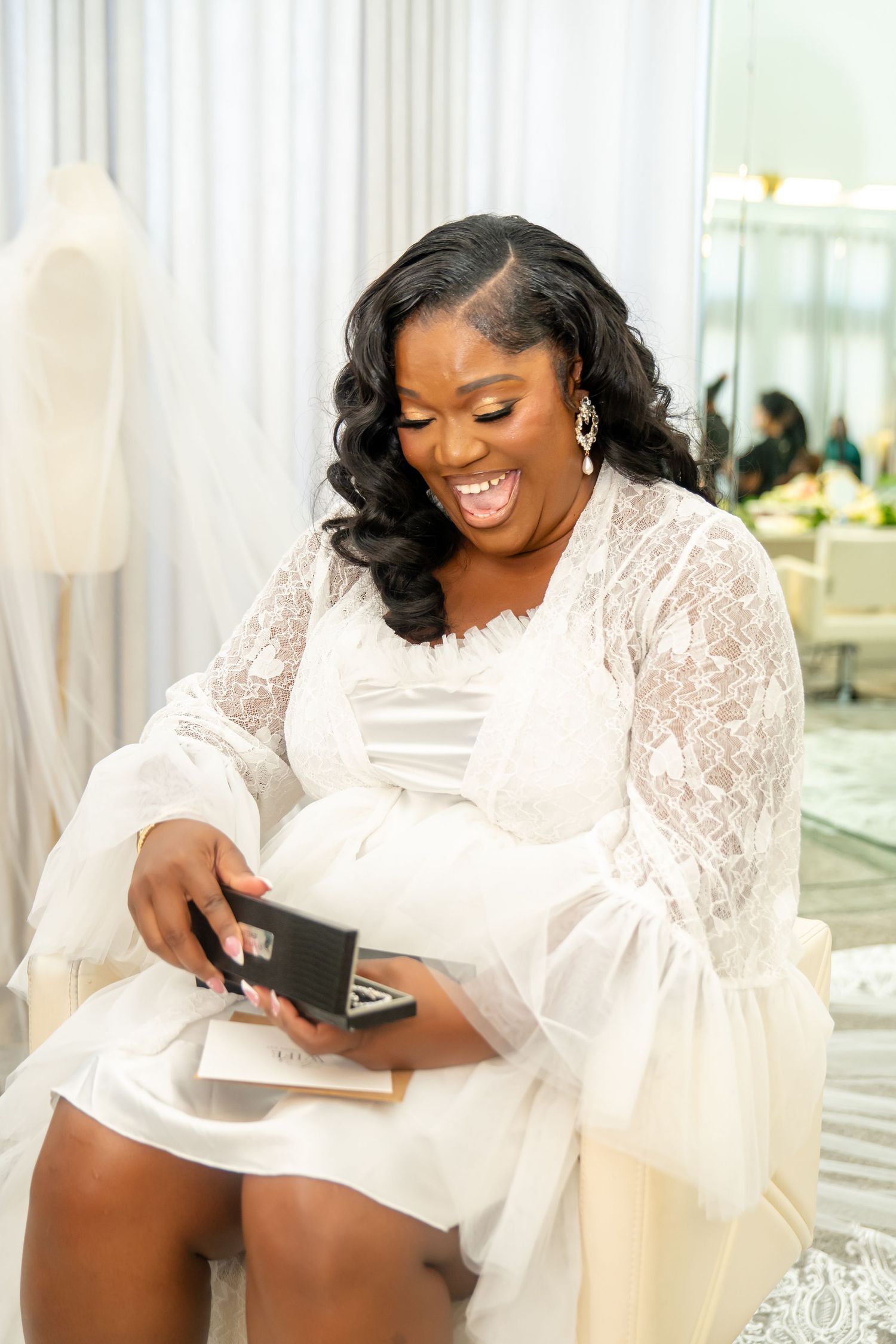 A person in white lace looks down with joy while holding a small box during wedding preparations.