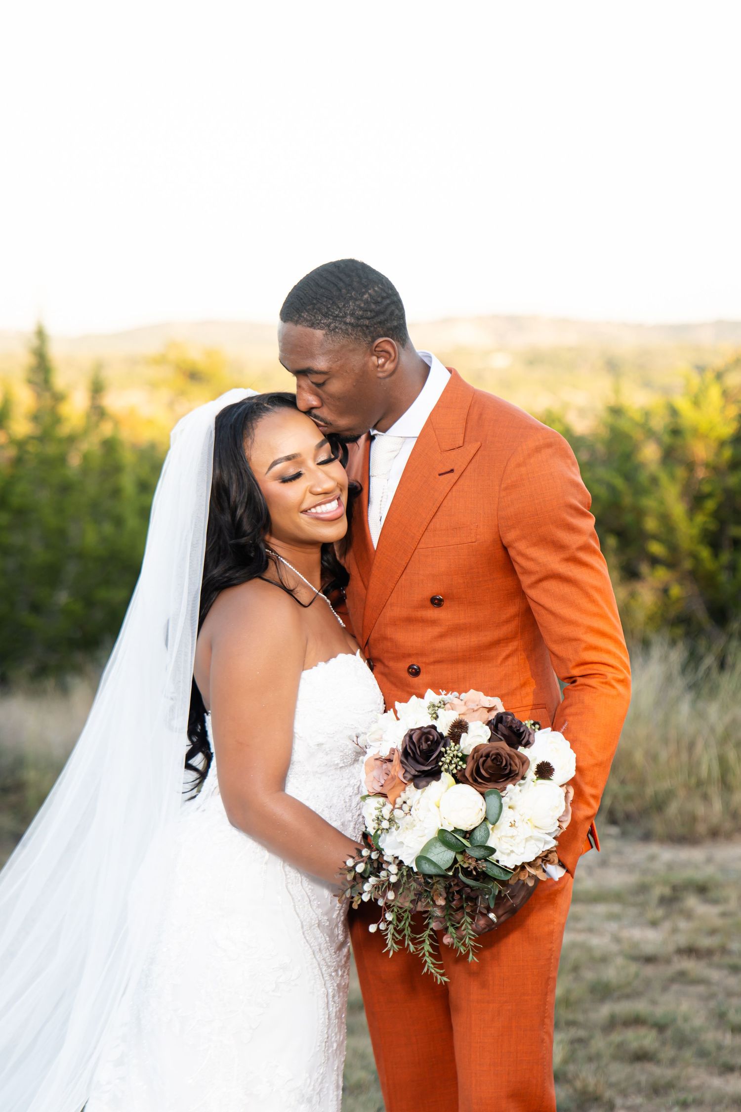 Wedding couple in white dress and orange suit embracing with bouquet outdoors.