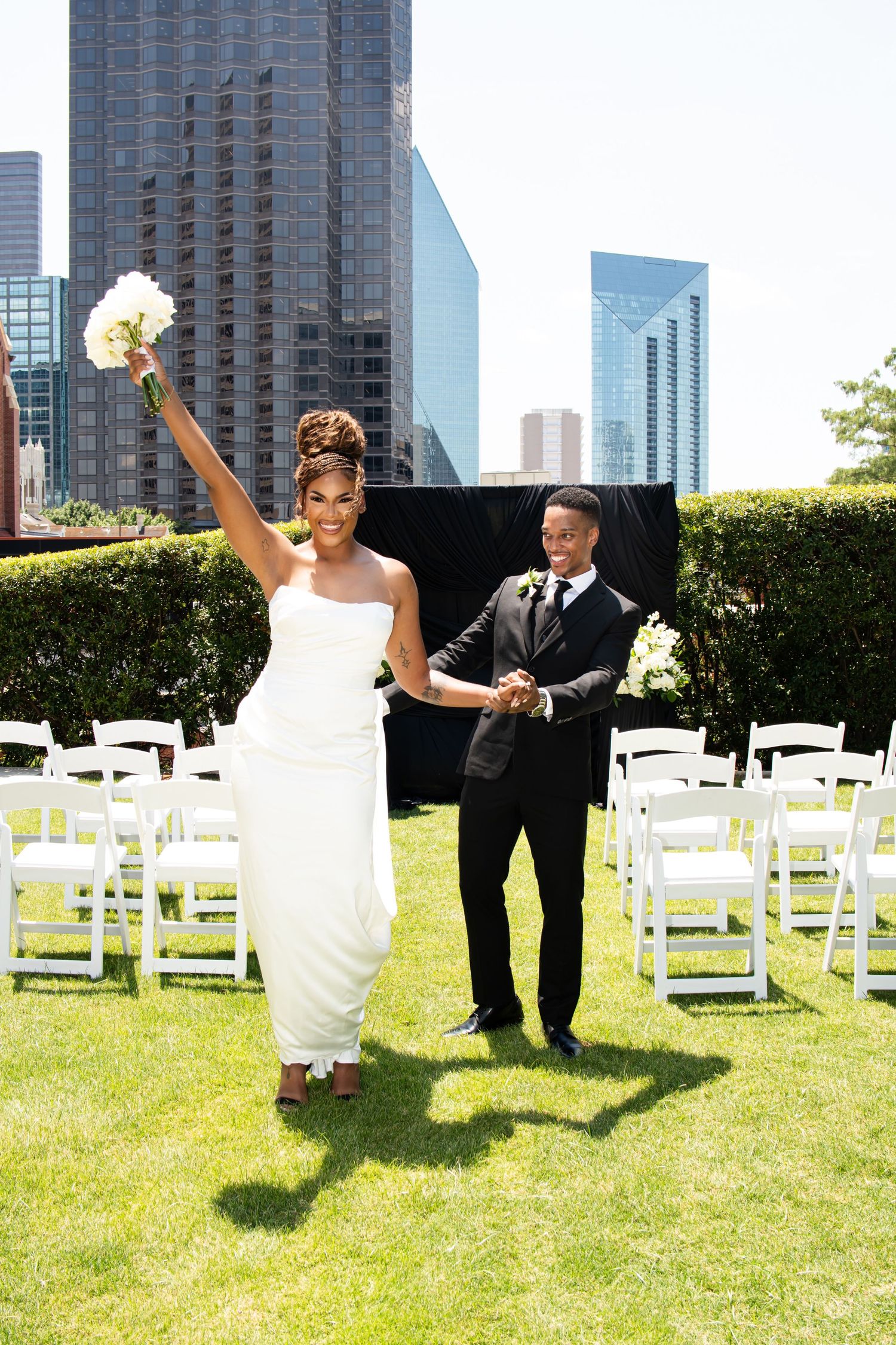 A joyful wedding celebration on a grassy lawn with white chairs and city buildings in the background.