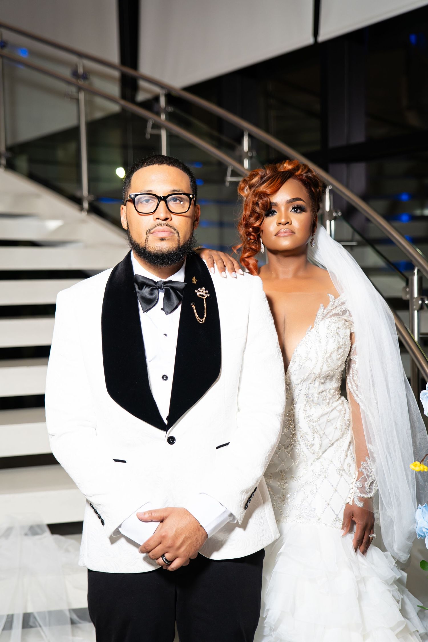 A wedding couple poses together in formal attire with the groom in a white tuxedo and bride in a lace wedding gown.