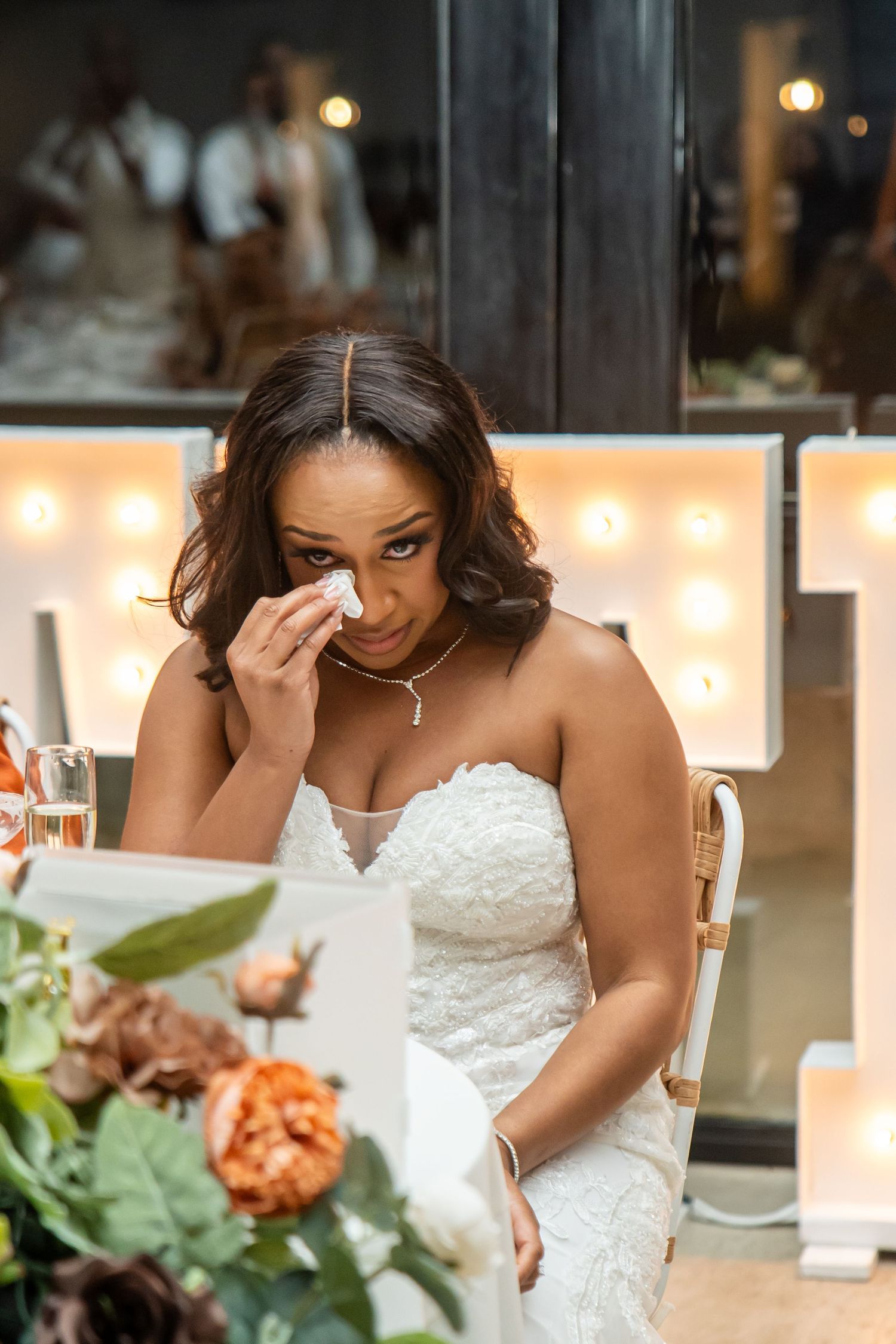 A person in a white lace wedding dress dabs their eyes with a tissue during an emotional moment.