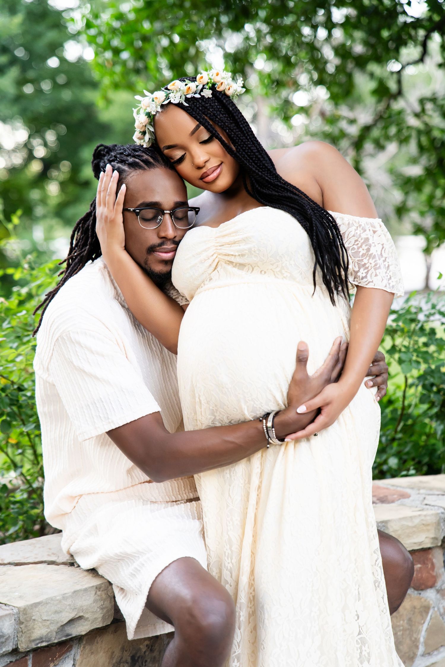 Intimate maternity portrait of couple in white outfits sitting on stone wall.