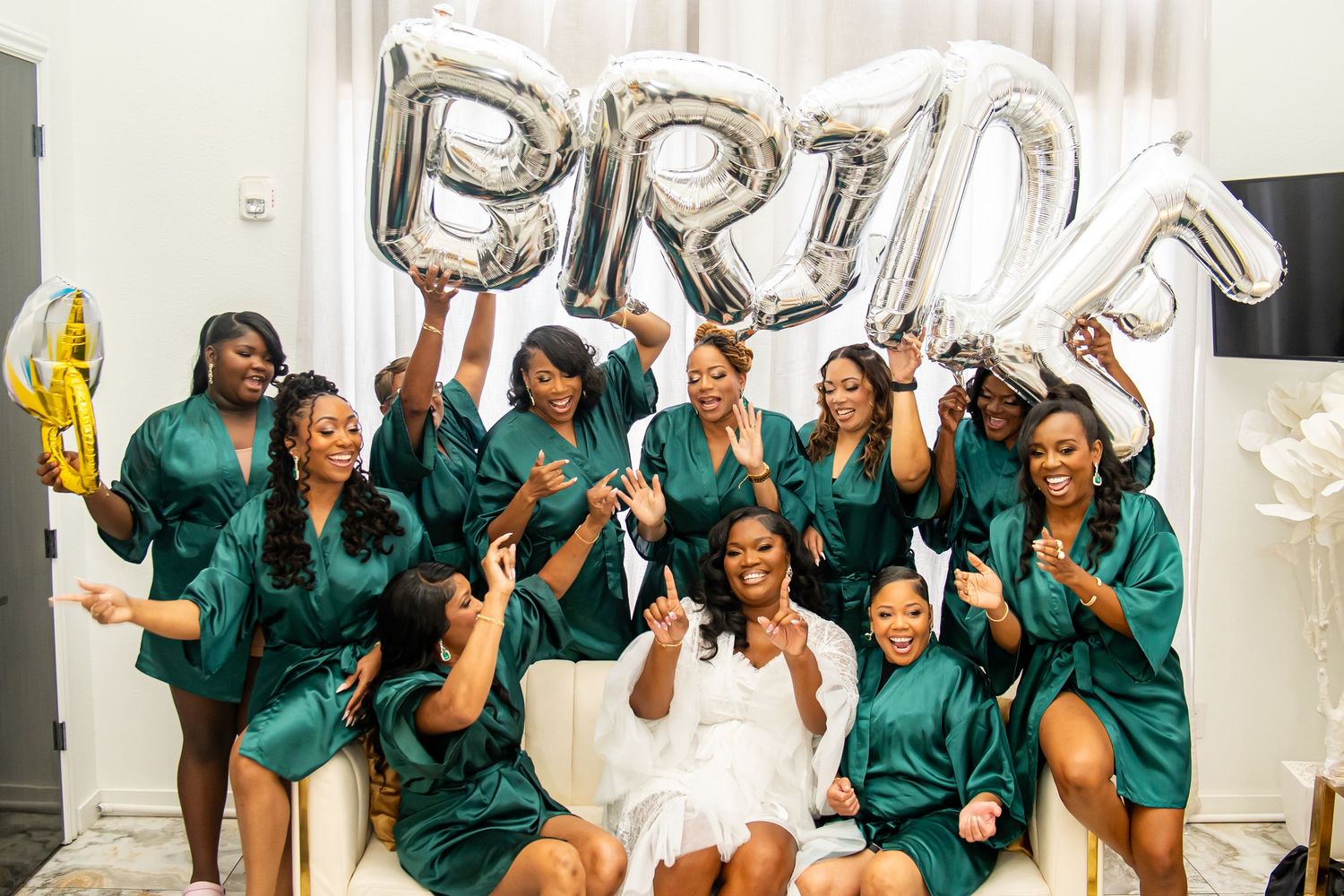 Bridesmaids in teal satin robes celebrate with silver letter balloons.