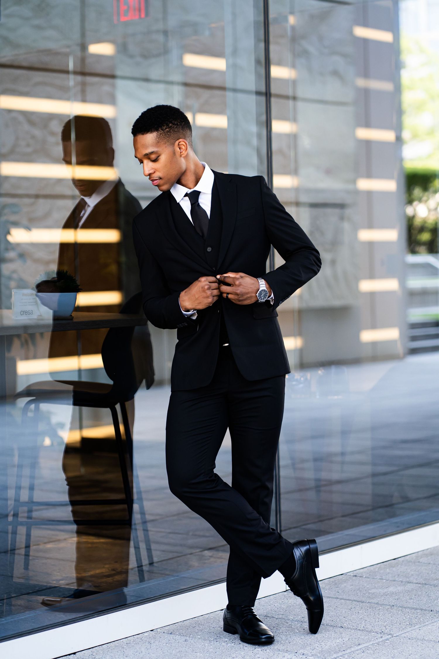 Person in a black suit adjusts their tie while standing against a modern building exterior.