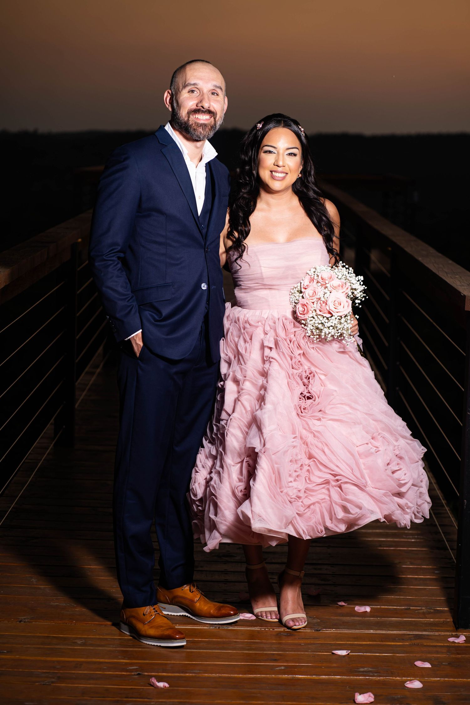 Couple poses at sunset, with bride in pink gown and groom in navy suit holding bouquet.