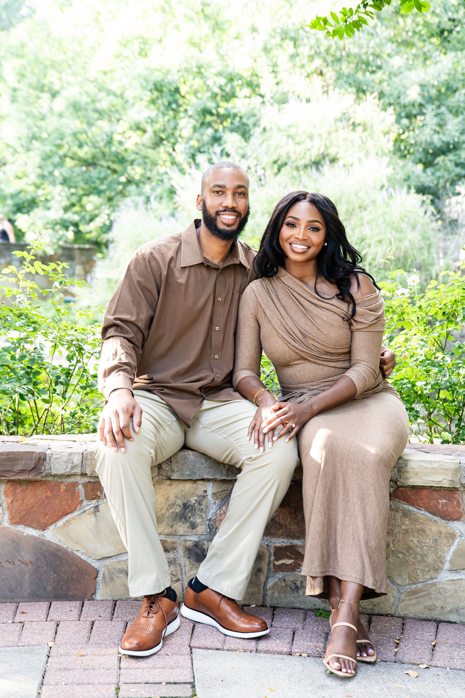Happy couple in coordinated brown and beige outfits sitting on stone wall outdoors.