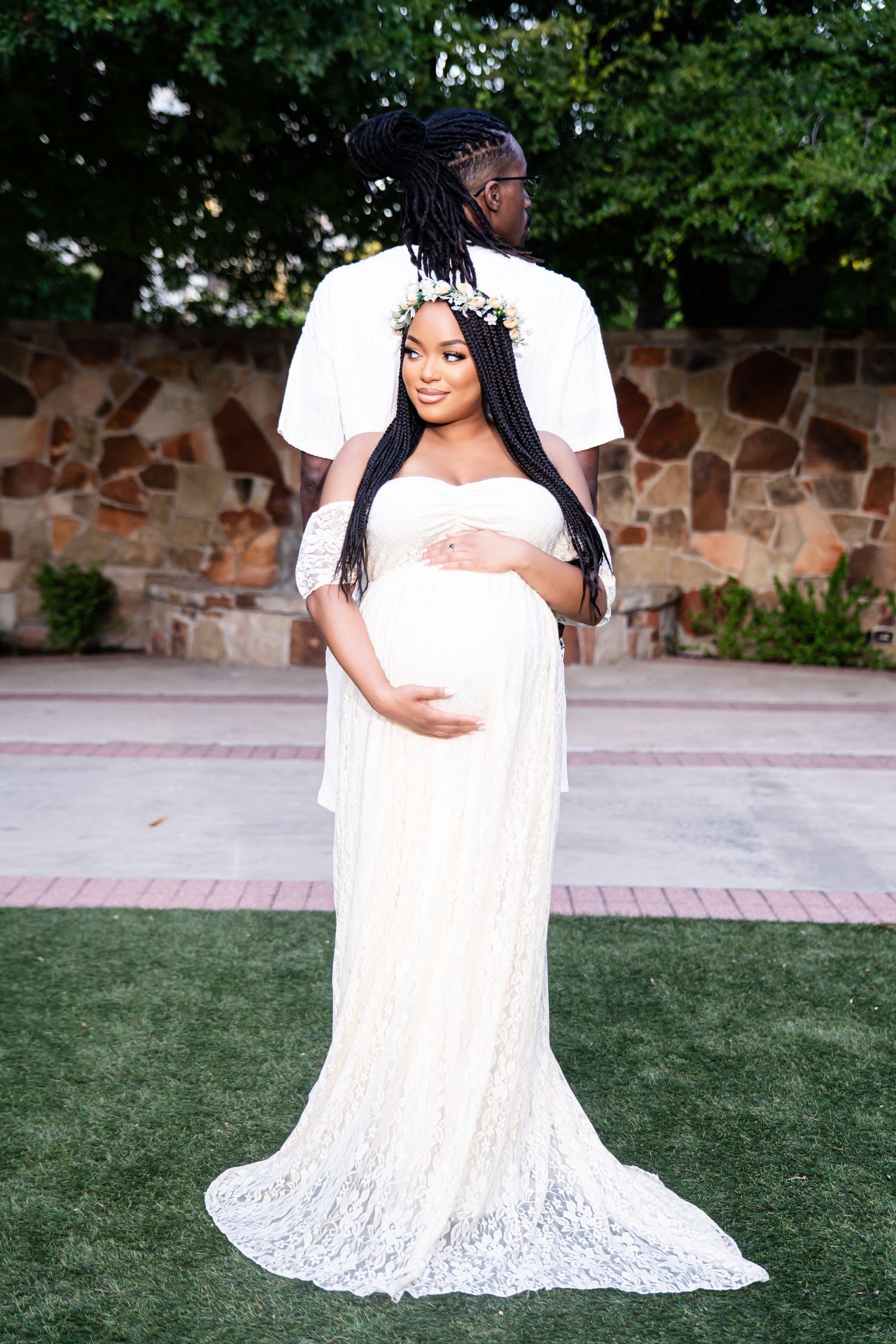 Elegant maternity photo featuring couple in white attire against stone wall backdrop.