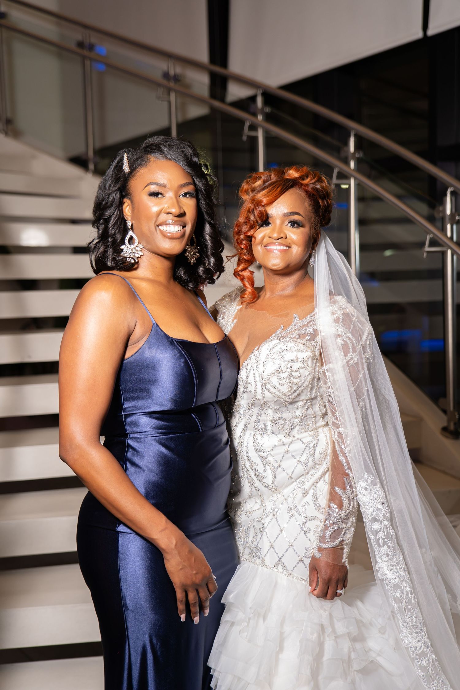 Joyful wedding party poses on stairs with bride in white gown and bridesmaid in navy dress.