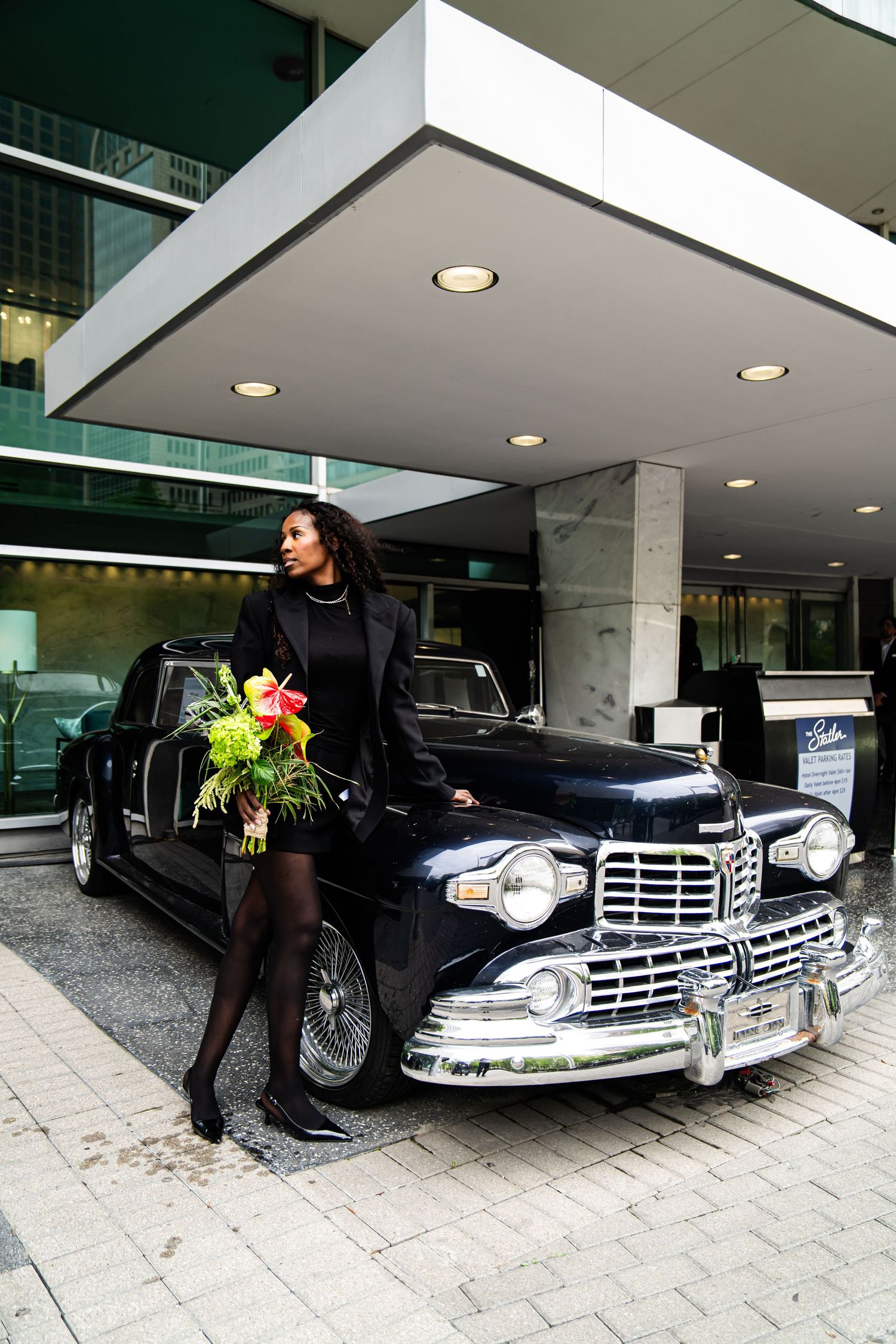 Classic black car displayed with model holding tropical flowers under modern building overhang.