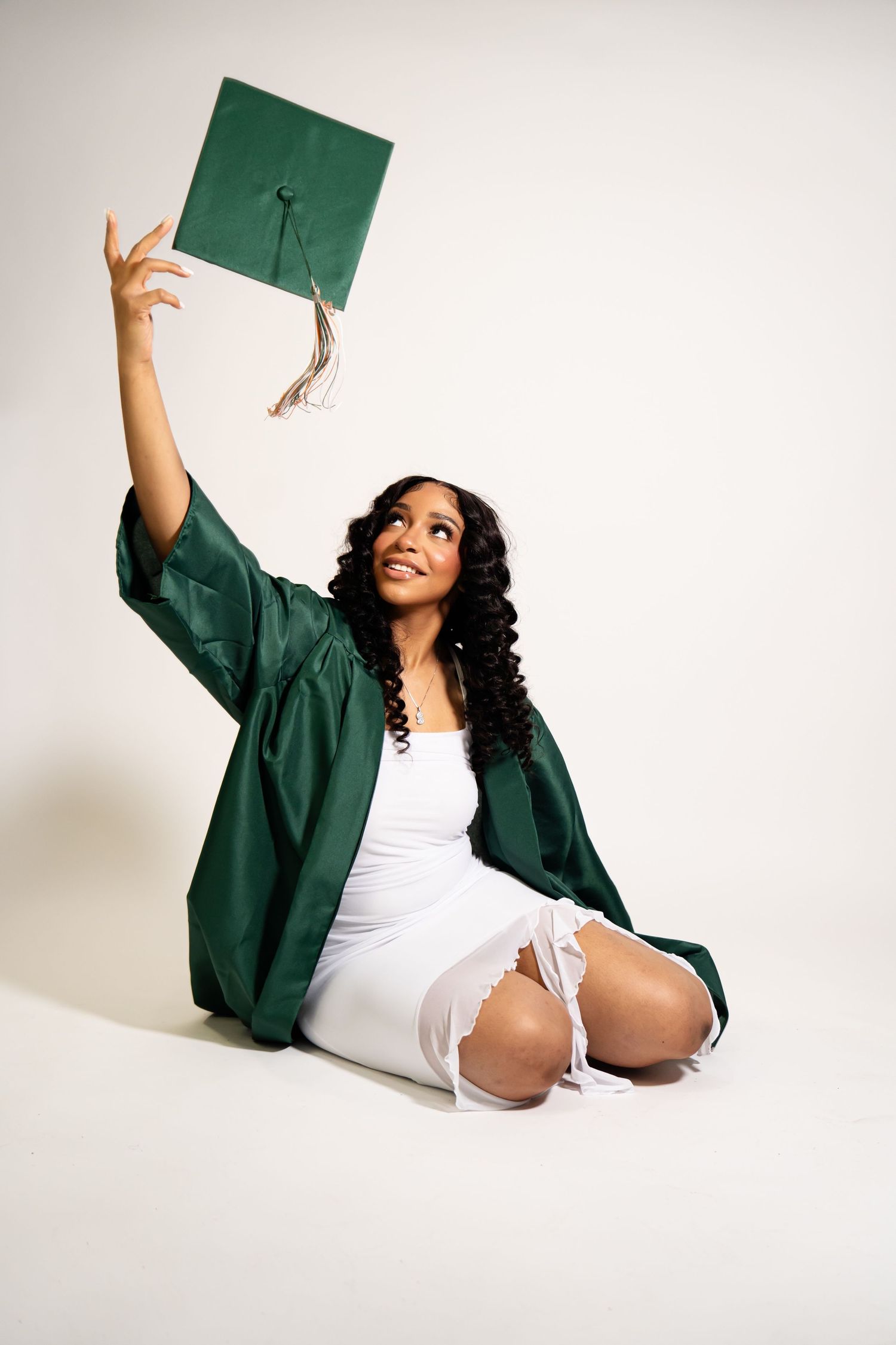 Graduate in green regalia strikes celebratory poses while sitting on white studio floor.