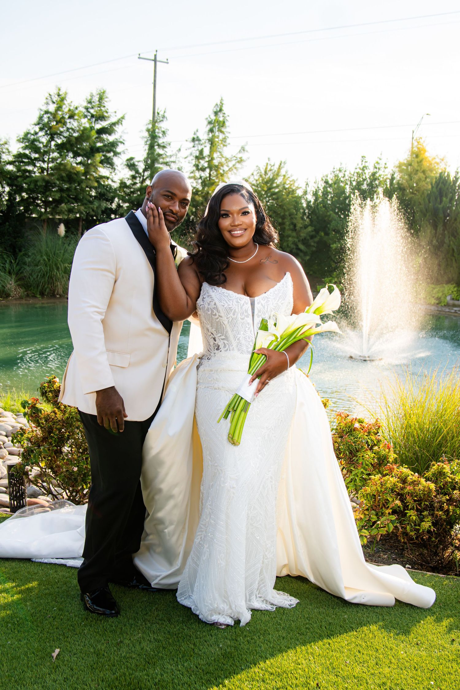 A couple poses for wedding photos in front of a fountain and garden landscape at sunset.