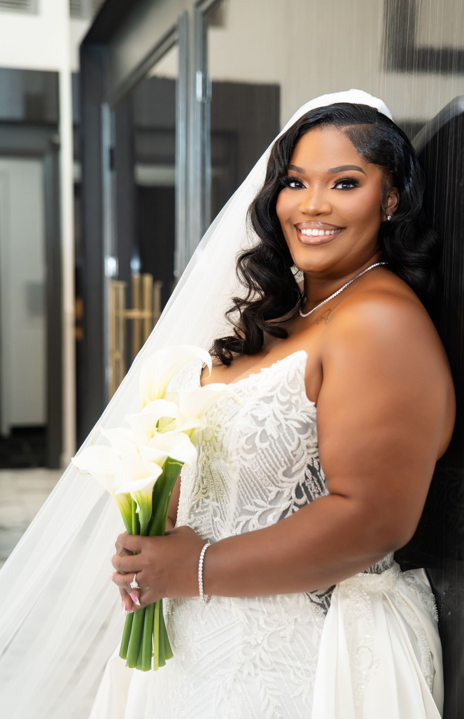 A person holds white calla lilies while wearing an elegant lace wedding gown with a long veil.