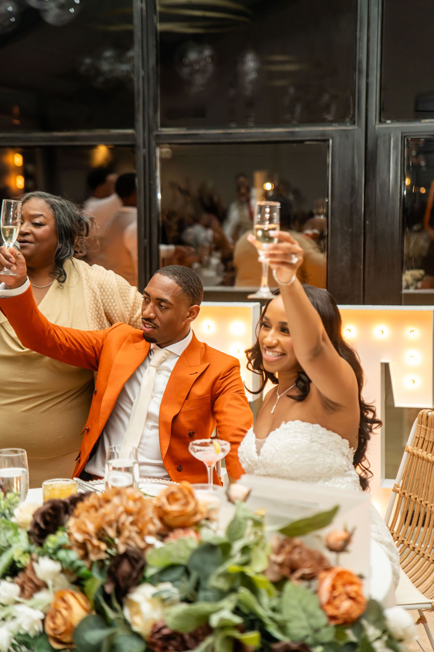 Wedding guests raise their glasses in celebration at a reception with decorative floral centerpieces and warm lighting.