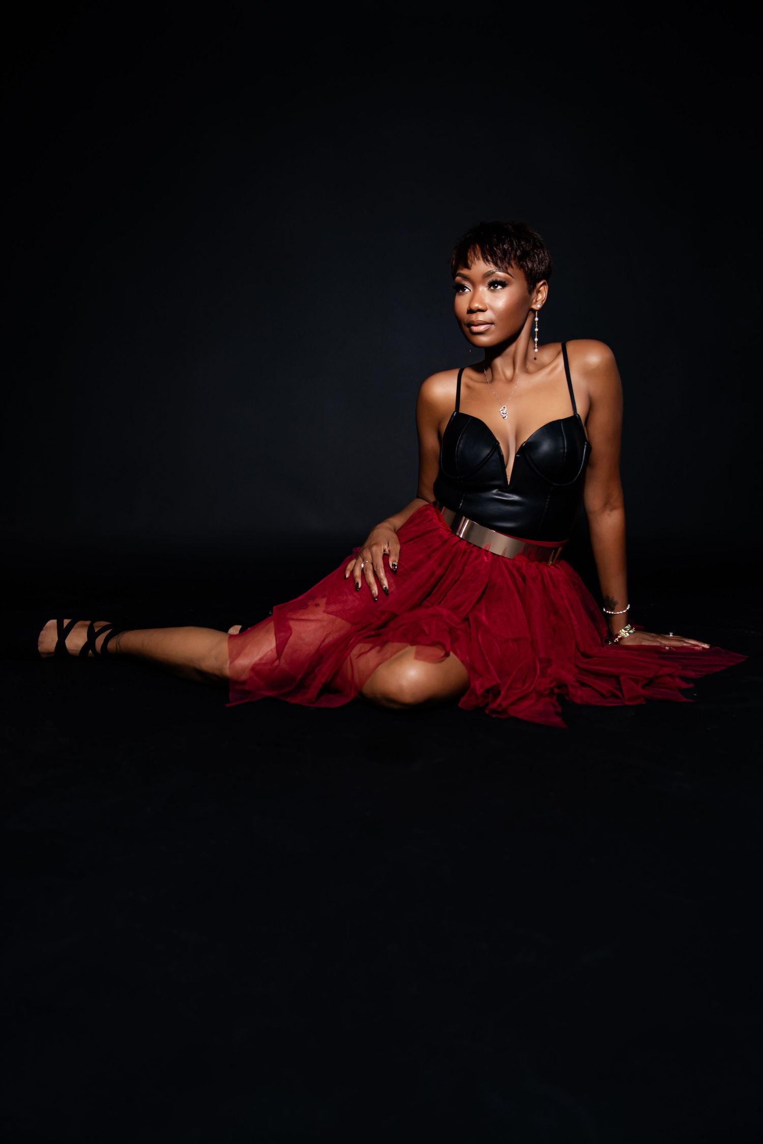 Fashion model in black bodysuit and flowing red dress poses dramatically against dark background.
