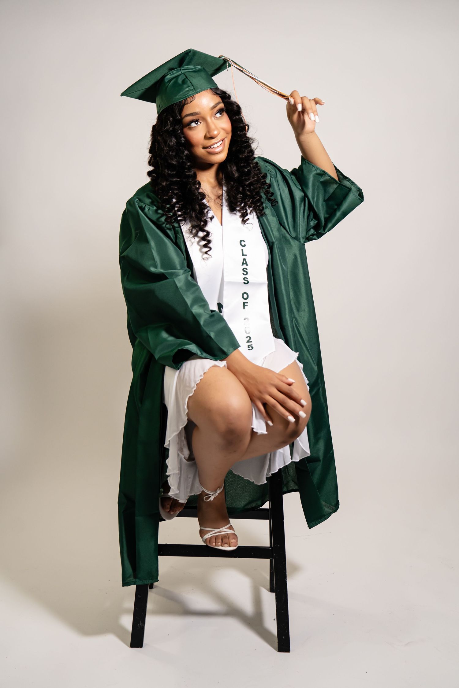 Graduate in emerald green cap and gown poses playfully on chair against white background.