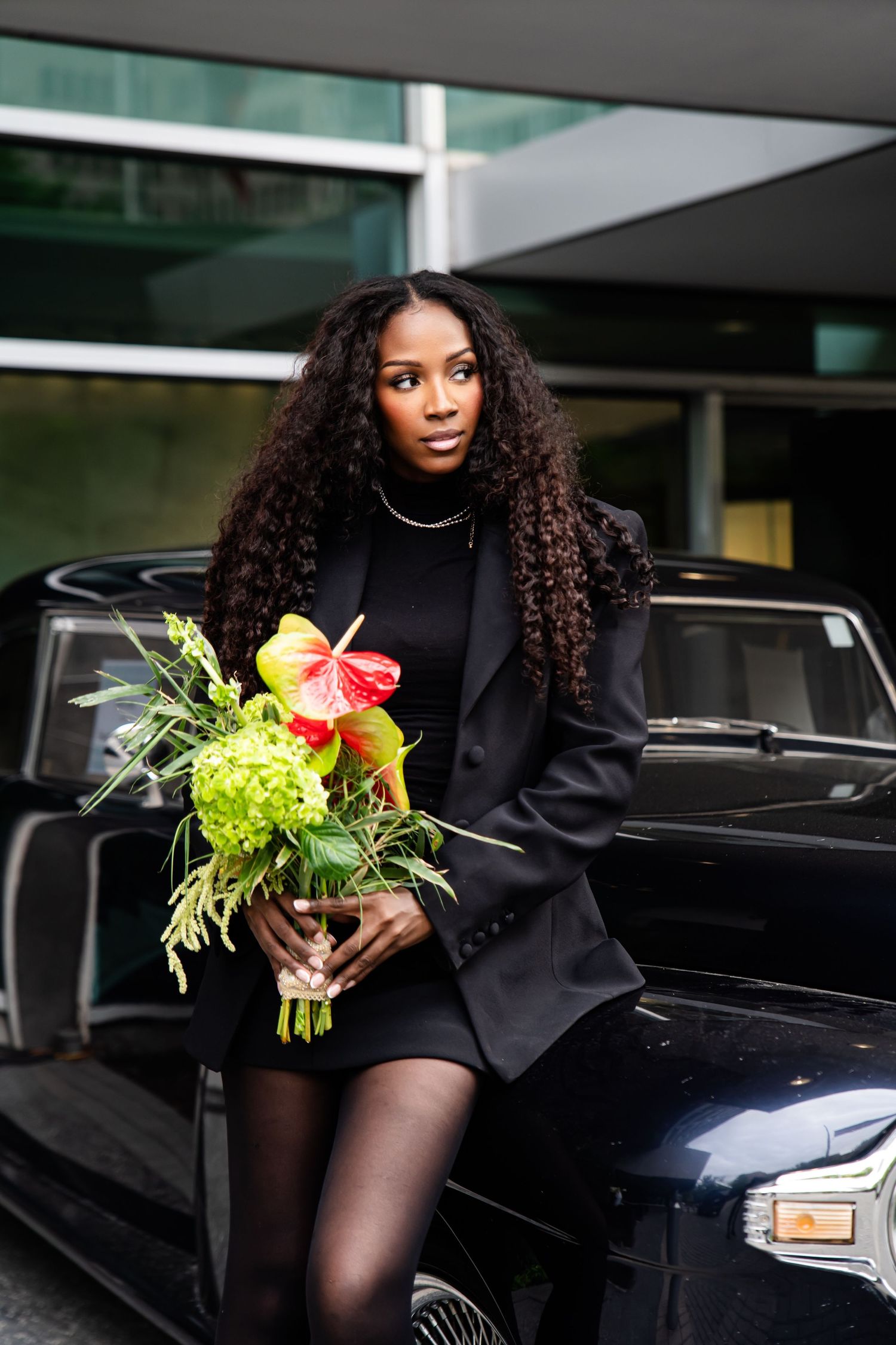Elegant model with bouquet poses beside vintage car in urban setting.