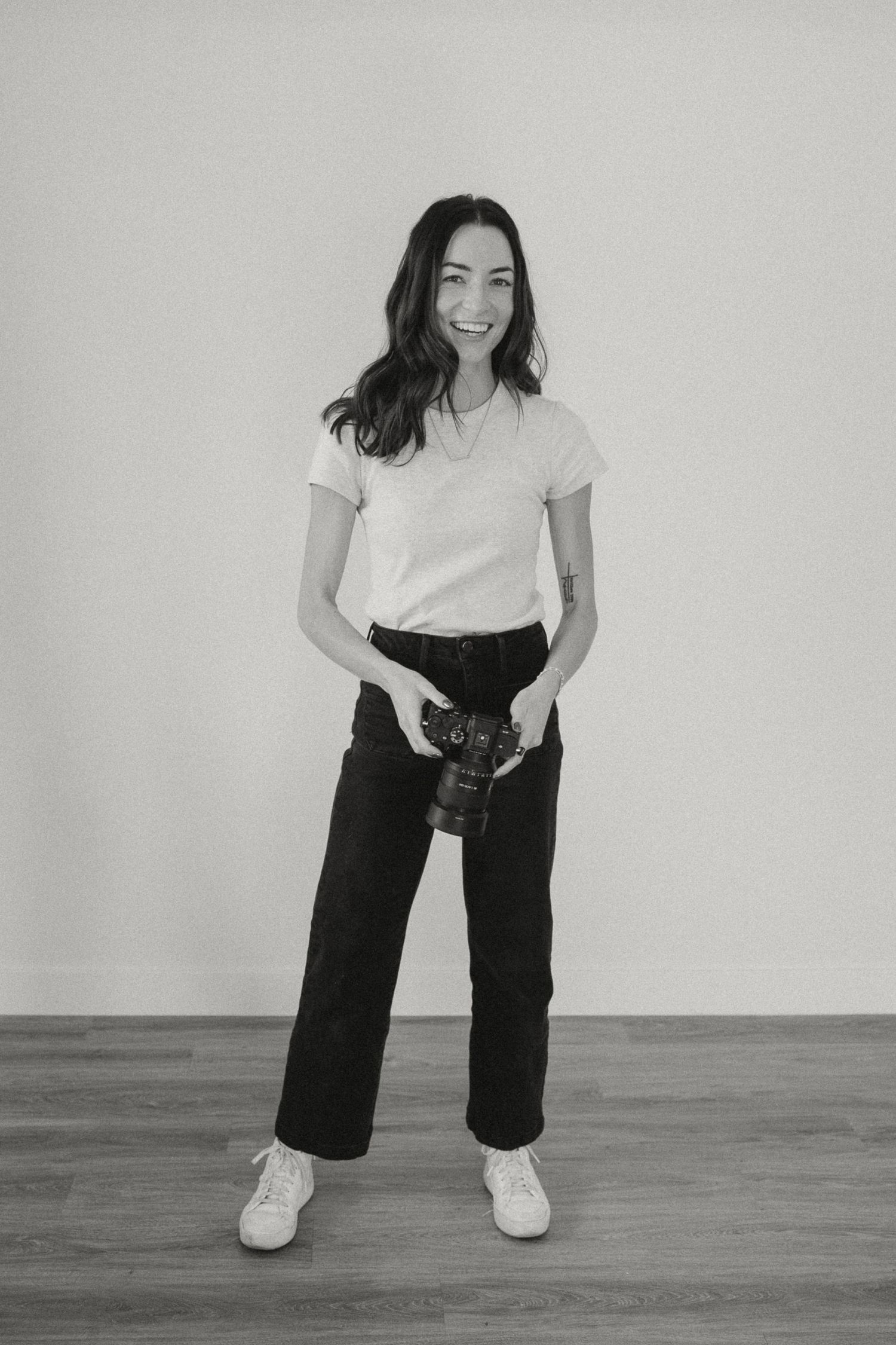 Professional photographer in casual attire holding a camera while standing against a white background.