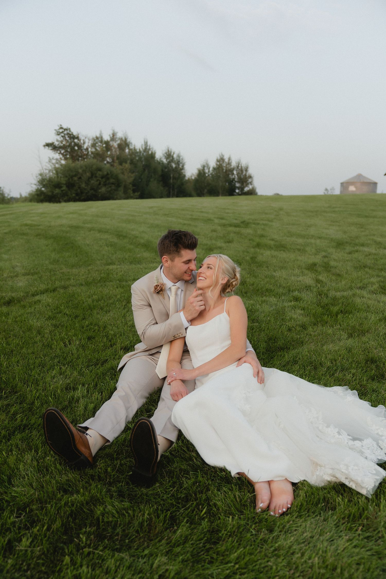 A couple shares a playful moment while lying in lush green grass during their wedding.