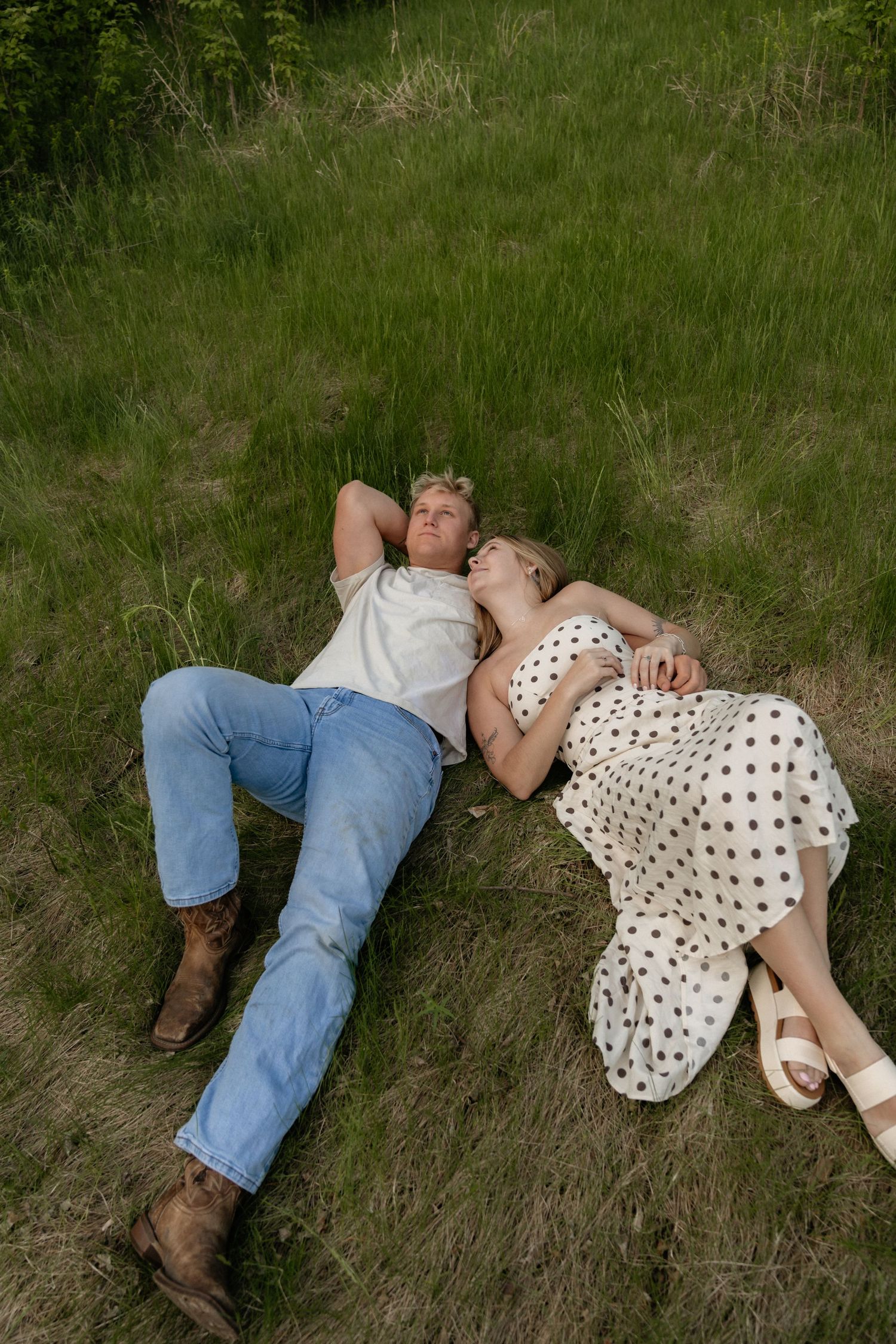 A couple lies together on green grass, woman in polka dot dress resting on man in jeans and cowboy boots.