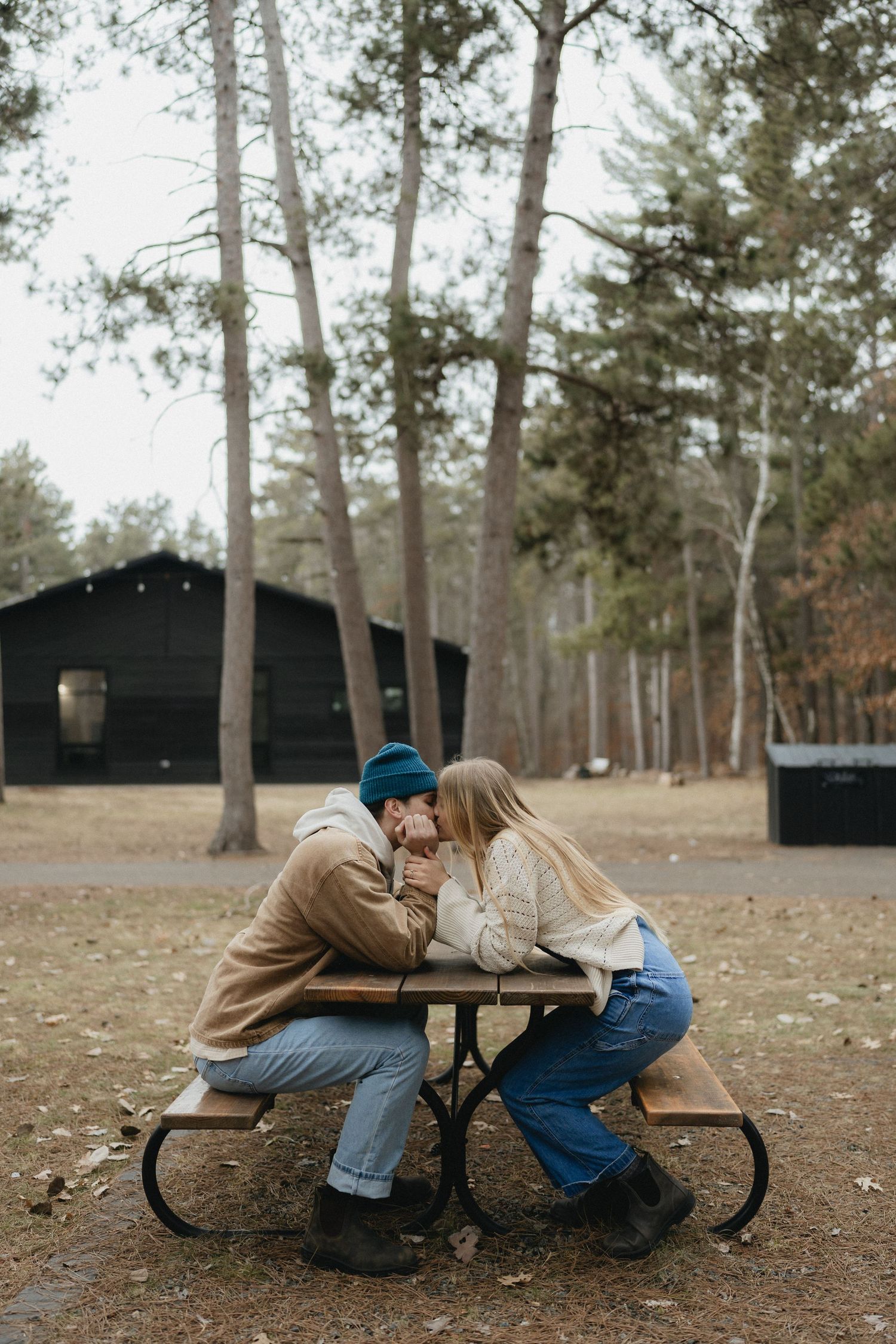 A couple shares an intimate moment at a wooden picnic table surrounded by tall pine trees.