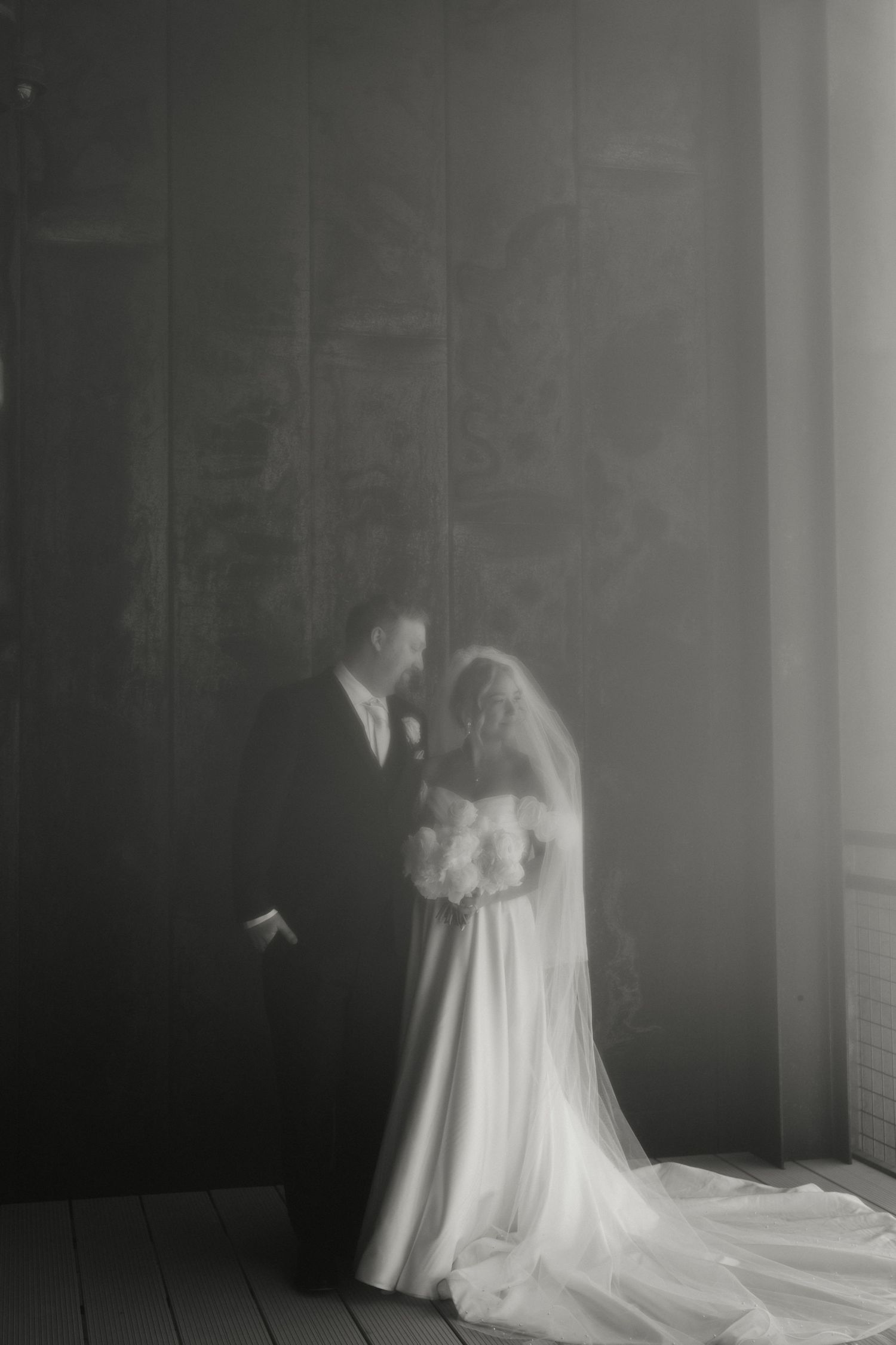 Black and white wedding photo of bride in veil holding bouquet and groom in suit sharing intimate moment by dark wall.