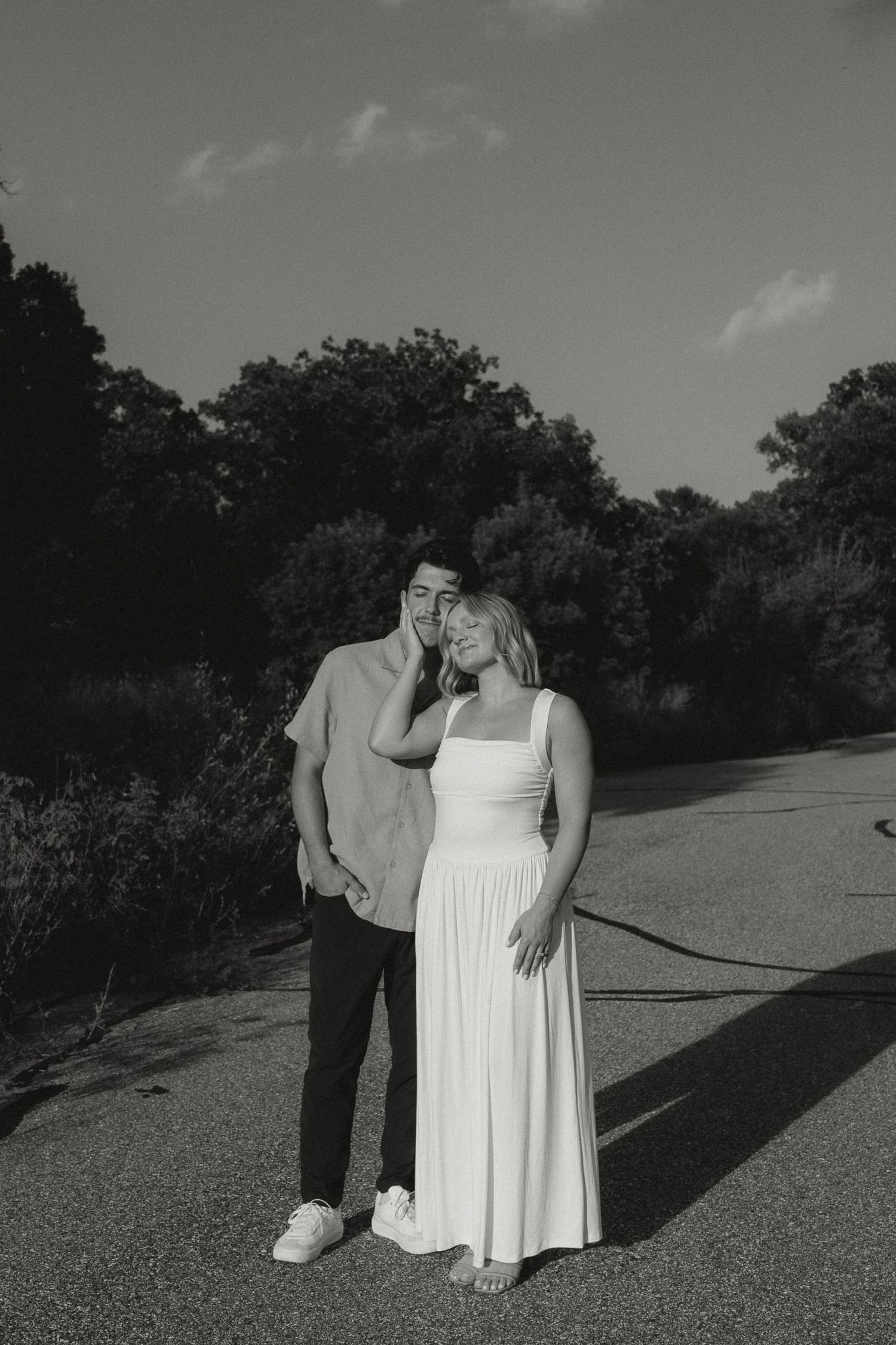 A couple shares an intimate moment on a gravel path surrounded by lush greenery in black and white.