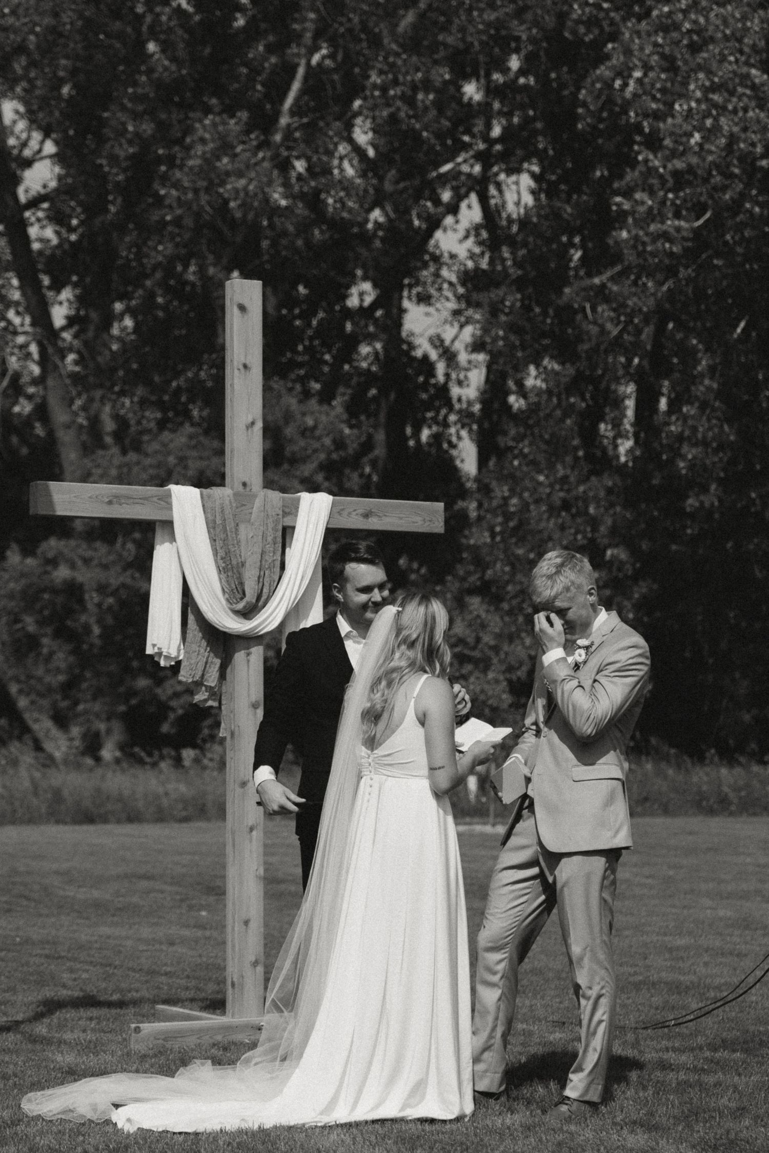 A wedding ceremony takes place outdoors with a wooden cross draped in white fabric.