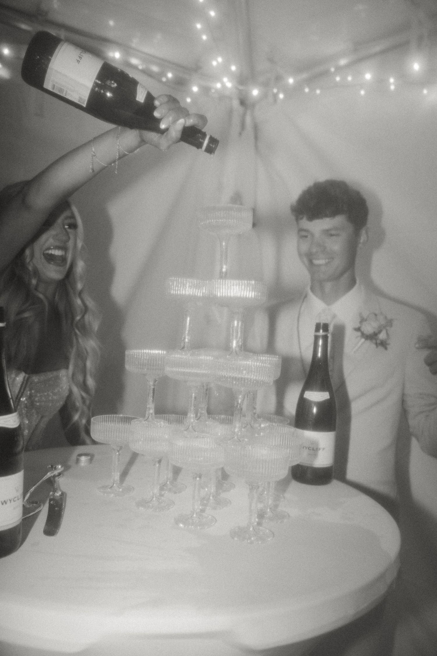 Wedding guests pour champagne into a tower of glasses under twinkling lights.