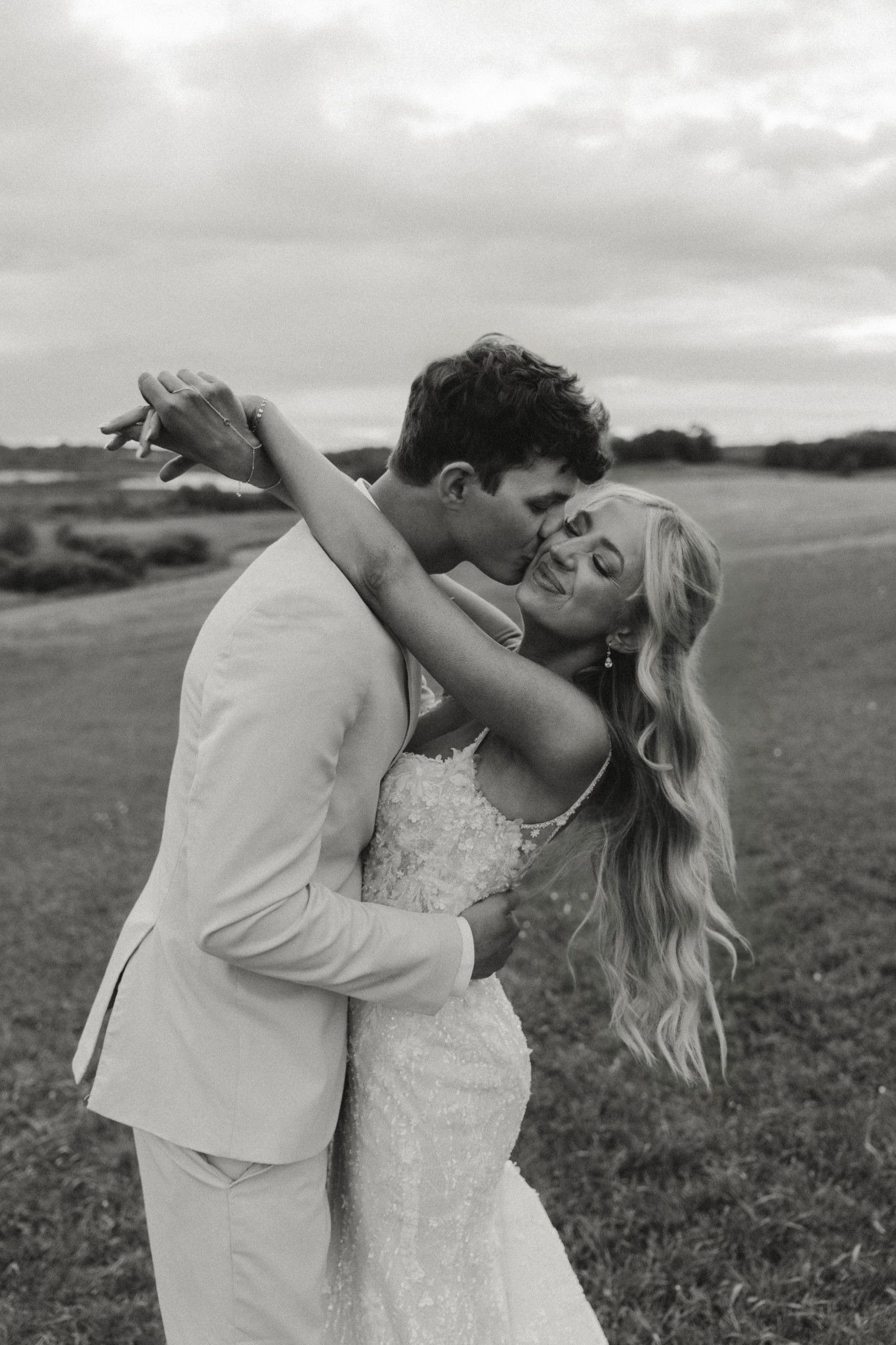Black and white photo of a groom kissing his bride on the cheek in a field on their wedding day.