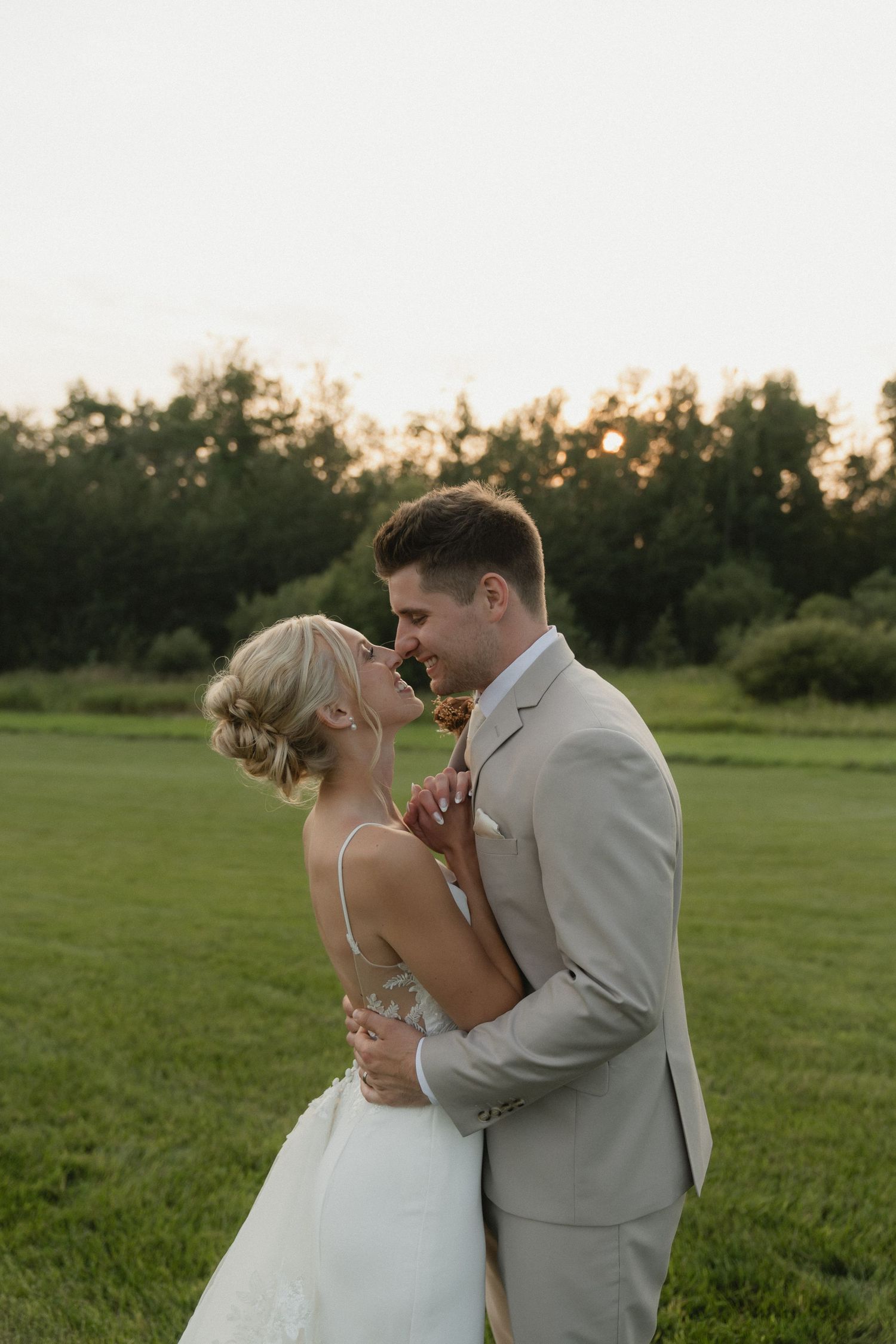 A bride and groom share a romantic moment at sunset in a lush green field, with the groom in a tan suit.