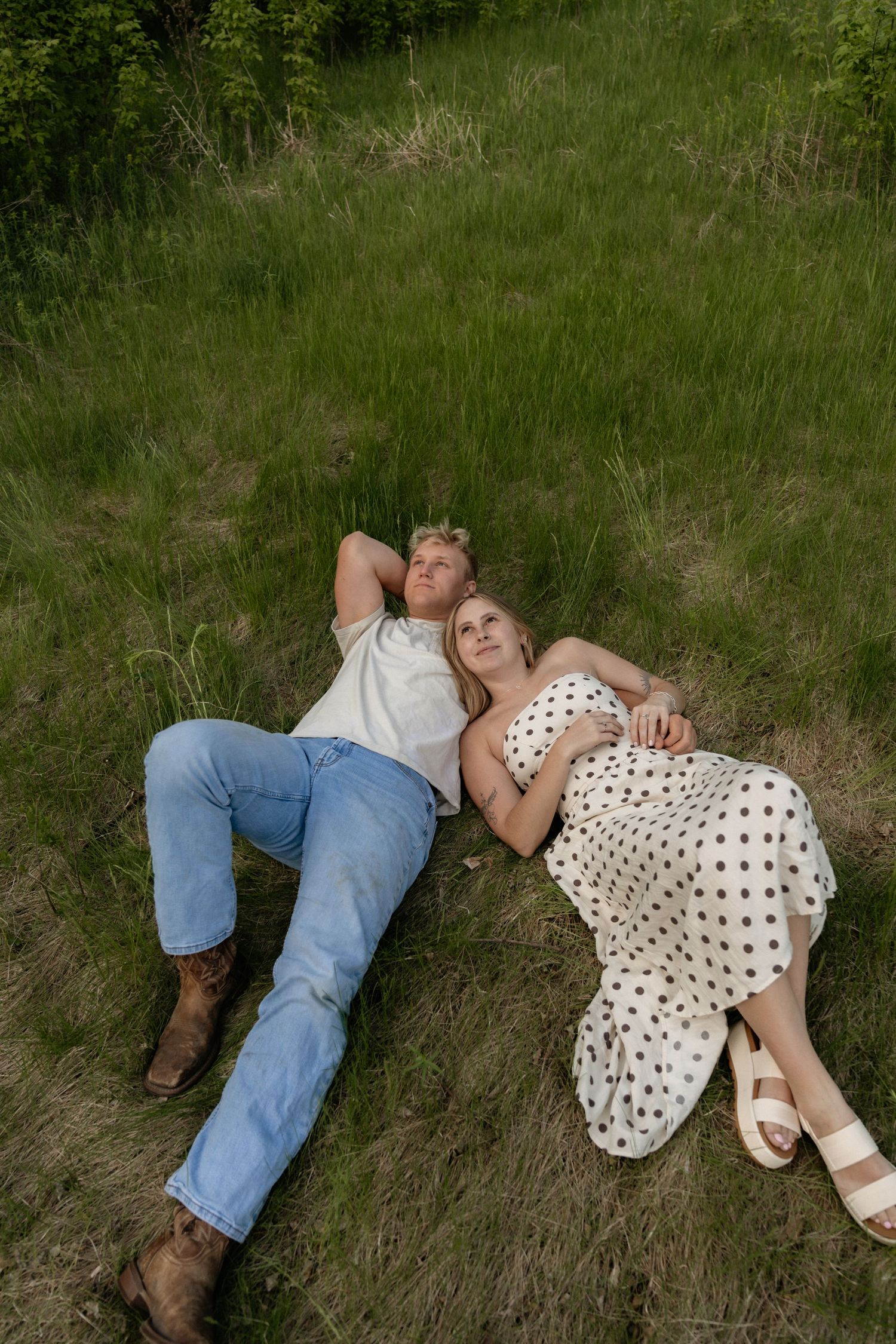 Couple lying on green grass hillside, woman in polka dot dress resting on man in jeans and white tee.
