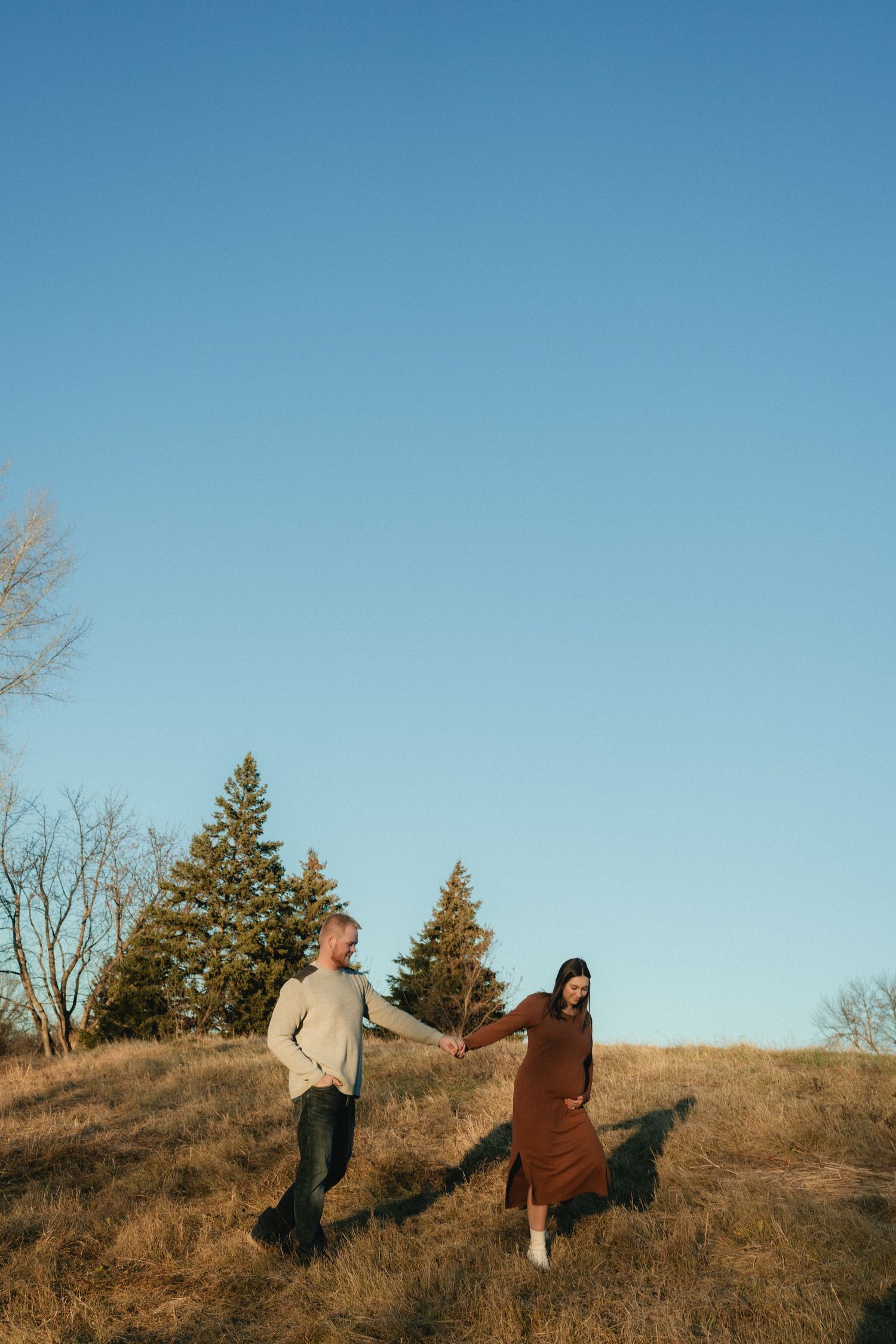 Couple holding hands walking on a golden hill with evergreen trees and clear blue sky in autumn.