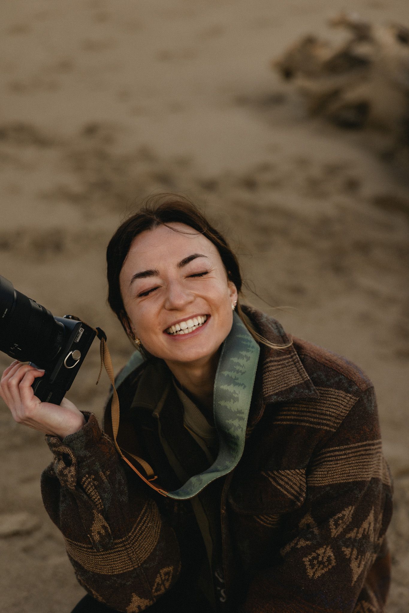 Smiling woman holding a camera outdoors on sandy beach, wearing a brown patterned coat with a teal scarf.
