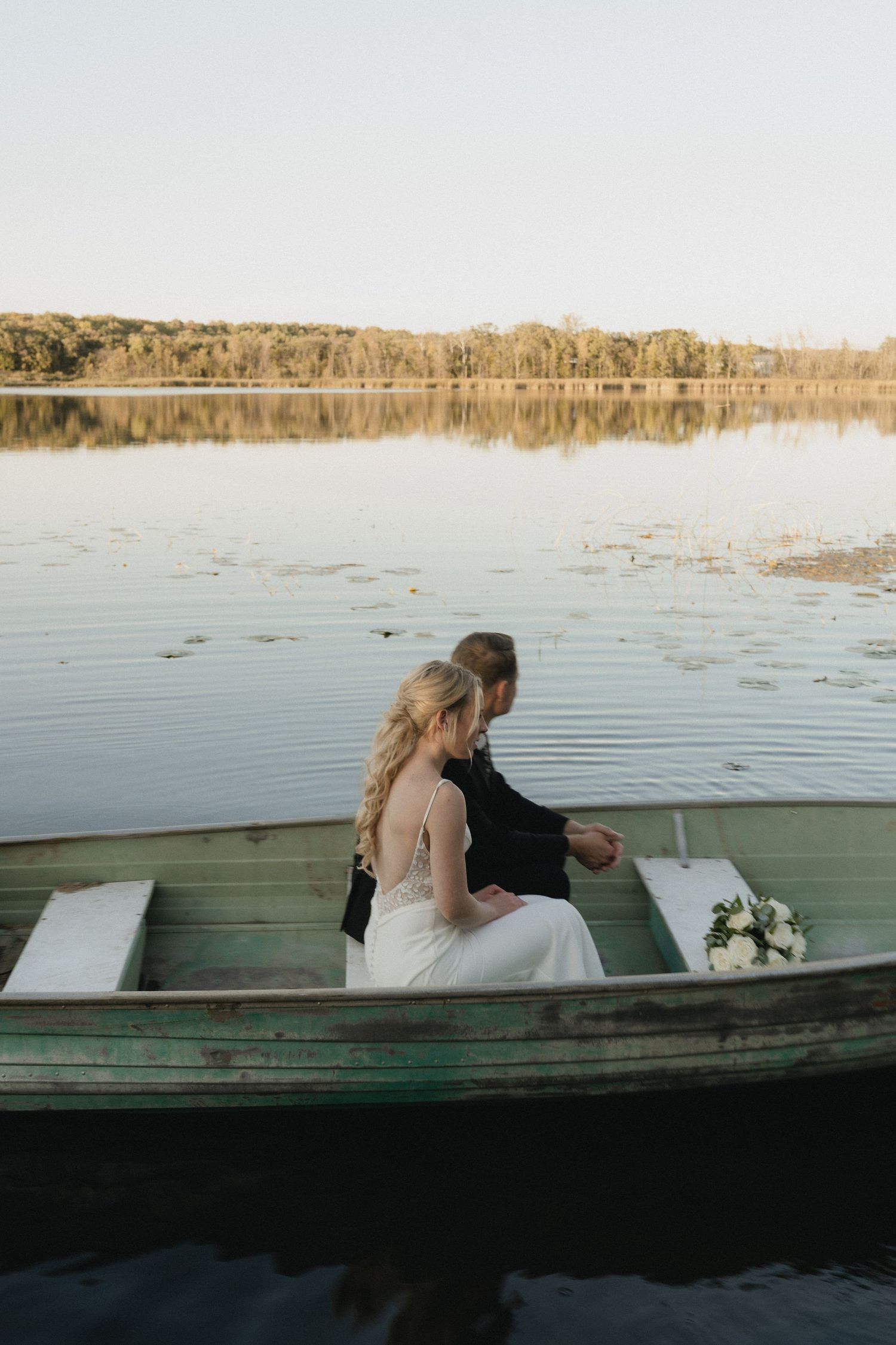 Bride and groom sitting together in a rustic green rowboat on a calm lake, with a white bouquet and tree reflections.