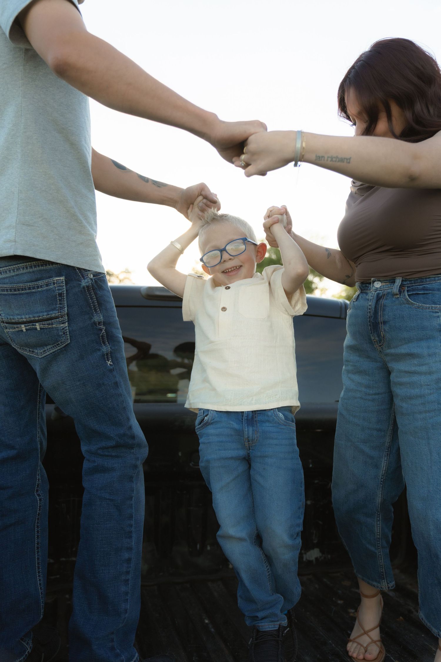 Parents holding hands with young boy wearing glasses outdoors, family bonding moment in casual denim outfits.