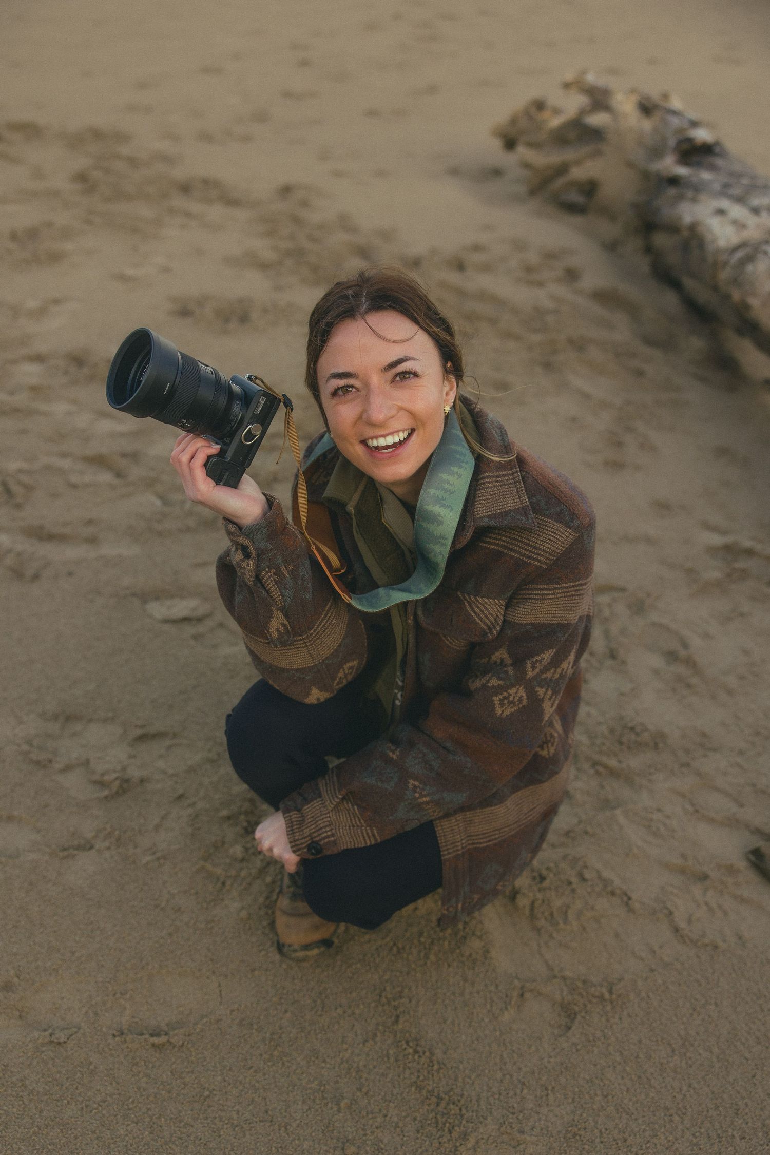 Photographer in brown jacket kneeling on beach sand with camera.