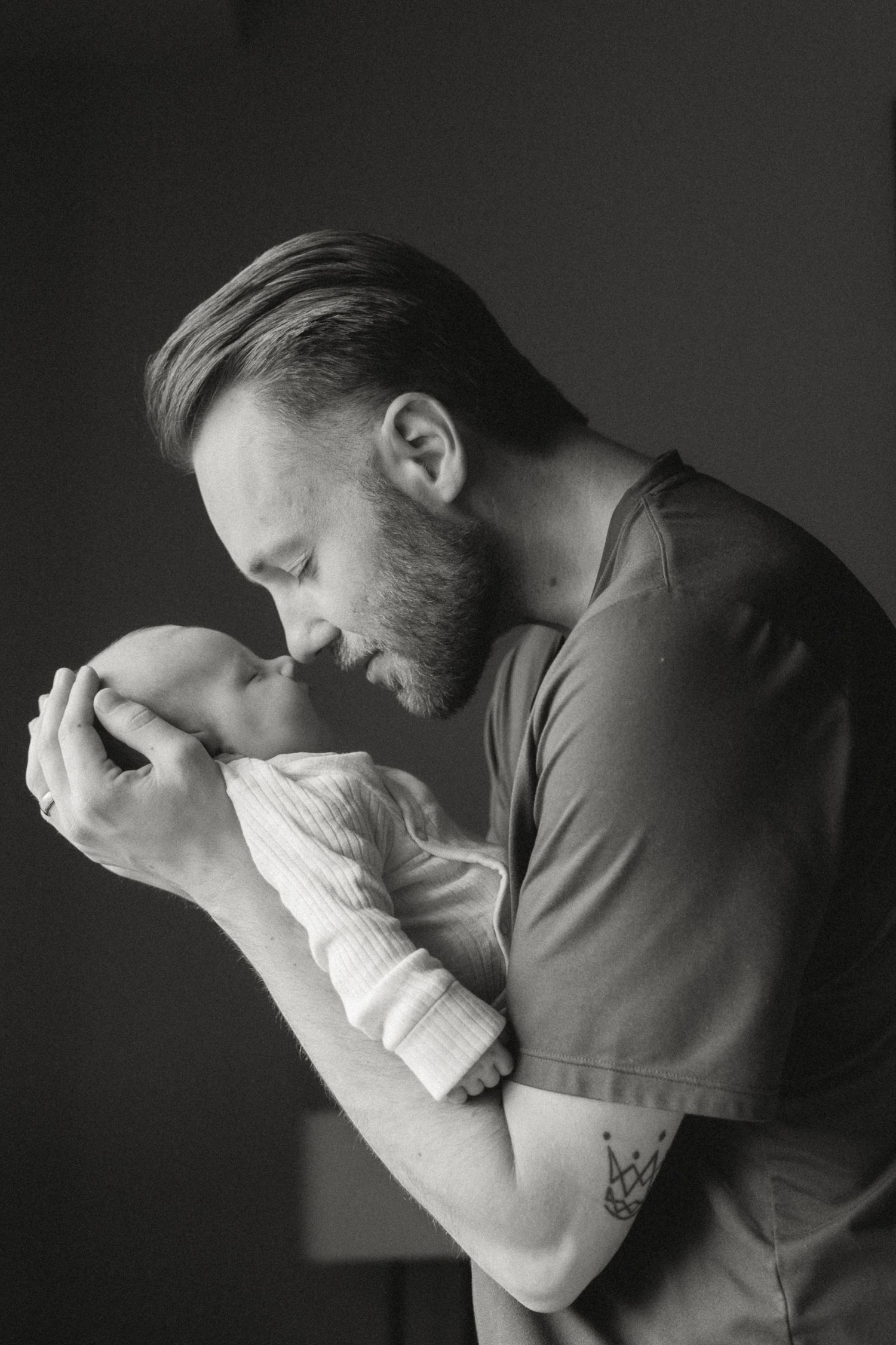 Black and white photo of a bearded father tenderly kissing his newborn baby wrapped in white cloth.